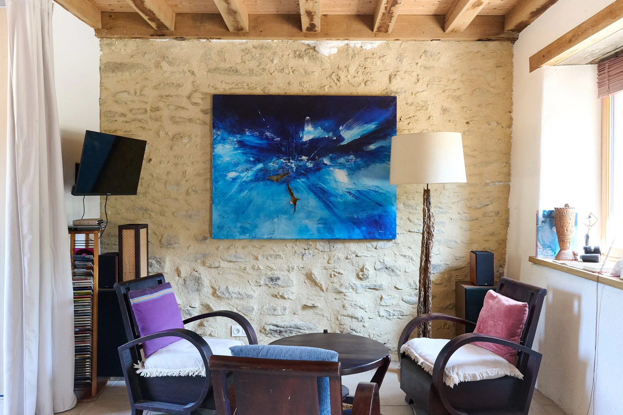 Living room with a brick wall, wooden ceiling beams, a large blue abstract painting, a flat screen TV, a white curtain, a floor lamp with a beige lampshade, and various chairs and decorative items.