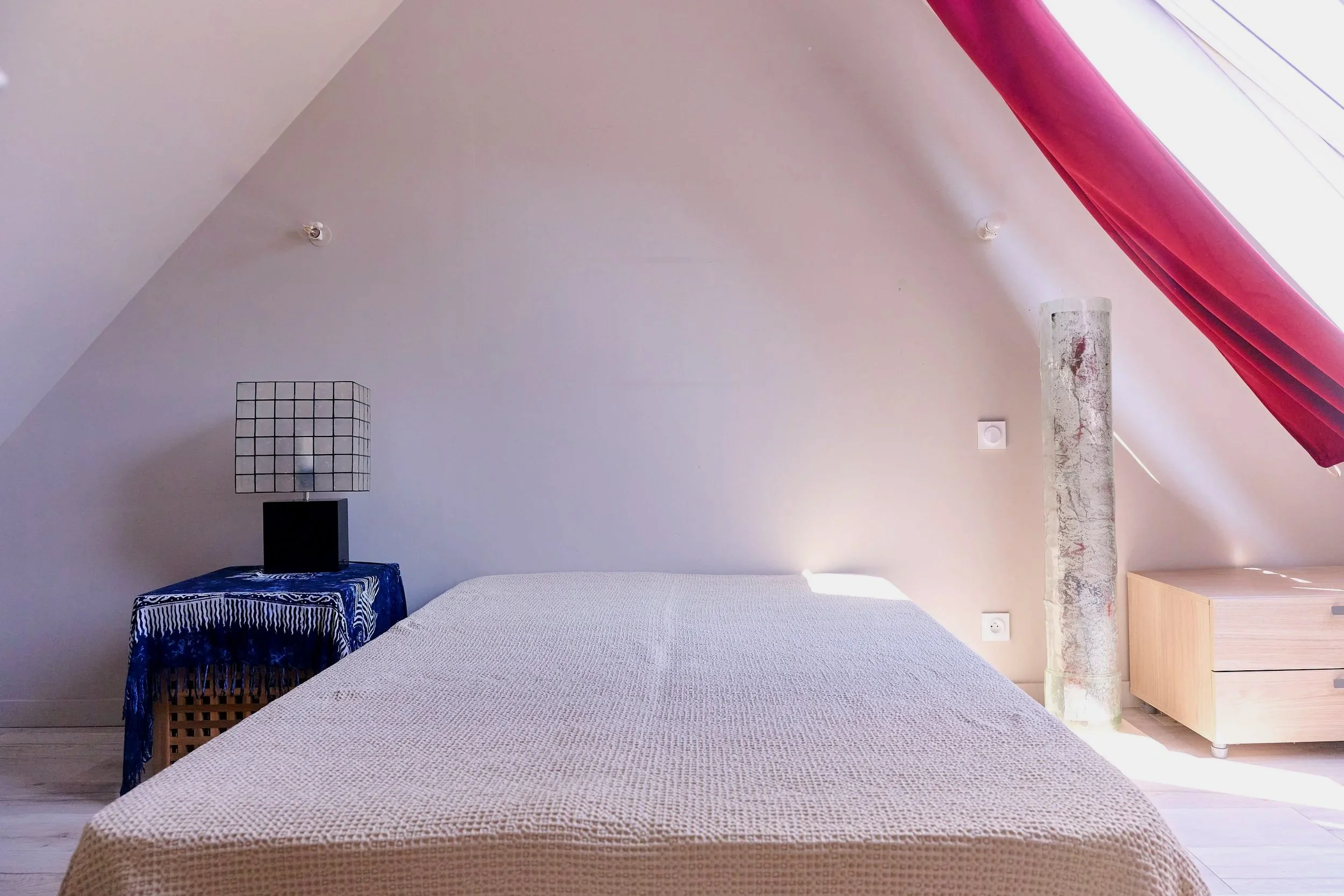Minimalist bedroom with a white bedspread, a black bedside table with a blue patterned cover, a lamp with a grid-patterned shade, a concrete pillar, and sloped ceiling with a pink beam and natural light.
