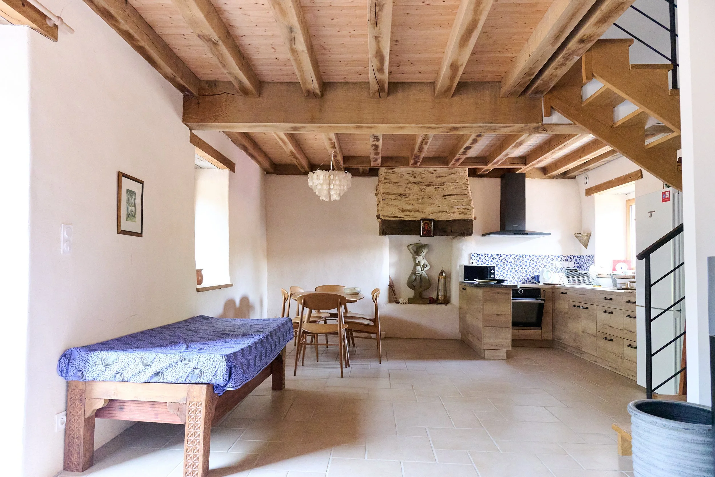 Interior of a room with a wooden ceiling with exposed beams, a chandelier, a stone fireplace with a religious icon, a kitchen area with wooden cabinets, a dining table with chairs, and framed artwork on the wall.