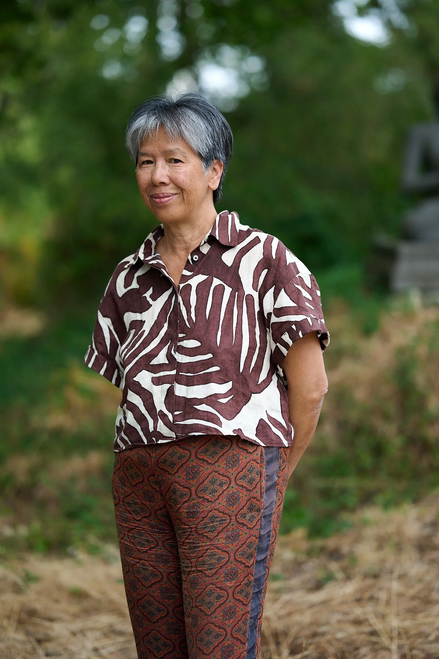 A woman with short gray hair standing outdoors in a green, blurred background. She is wearing a patterned shirt with brown and white designs and patterned pants. She is smiling softly and positioned slightly turned to her left.