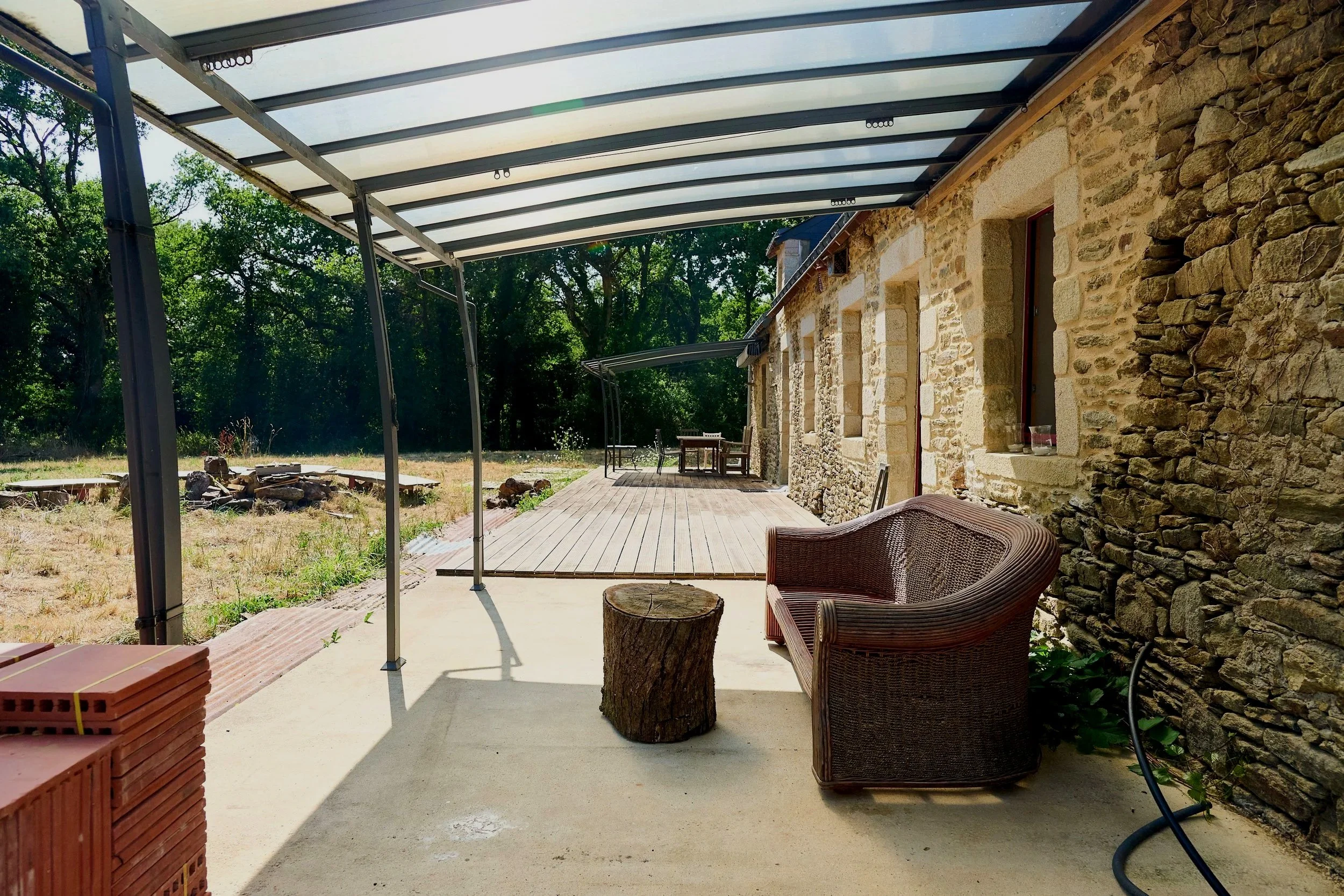 The image shows a covered outdoor area attached to a stone house, with wooden decking flooring, wicker chairs, a tree stump used as a table, and a stack of bricks. The roof is made of transparent panels supported by a metal frame, and there are trees and greenery in the background.
