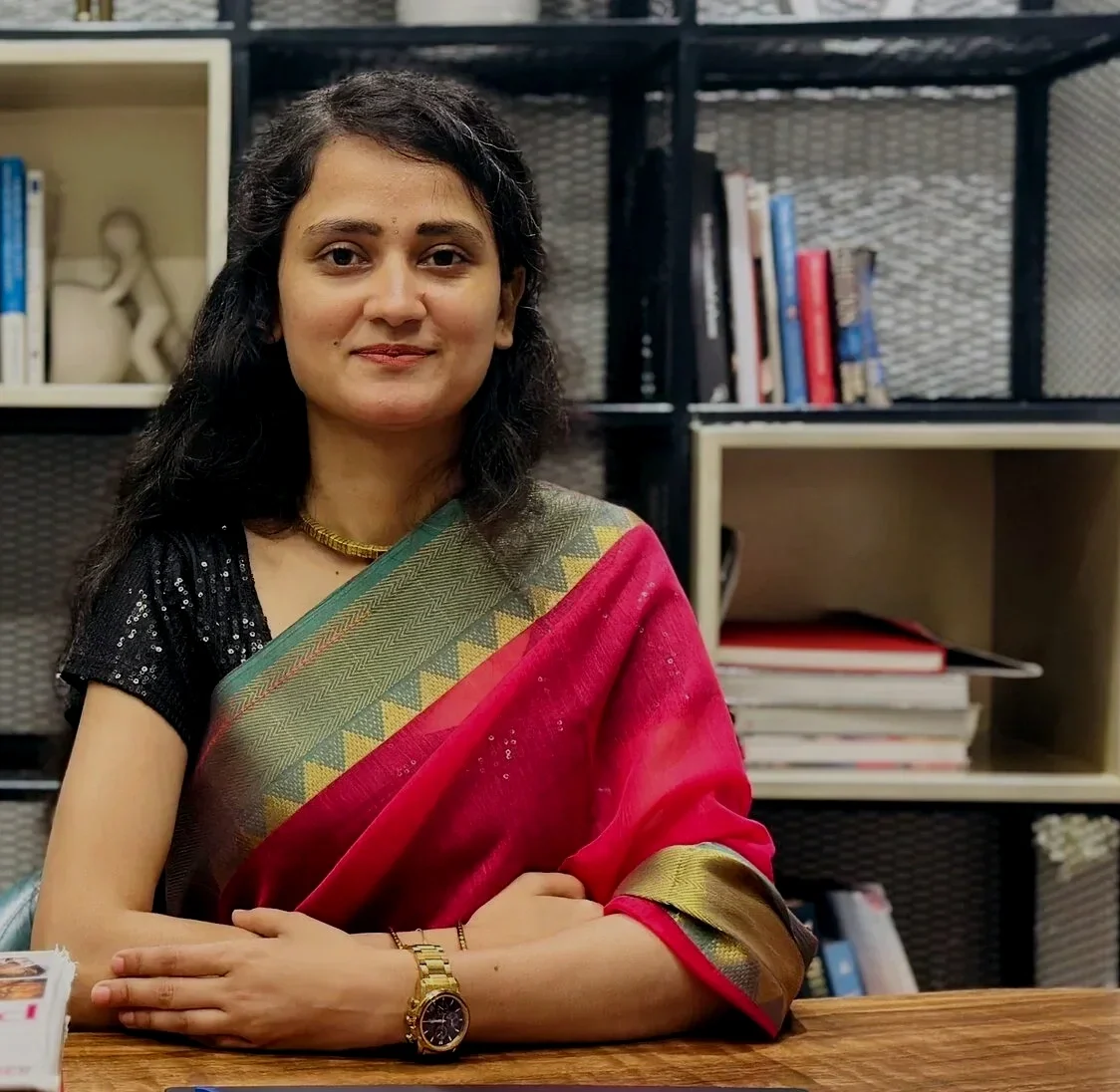 A woman with dark hair wearing a saree with red, green, and yellow patterns, sitting at a wooden table, in front of shelves filled with books.