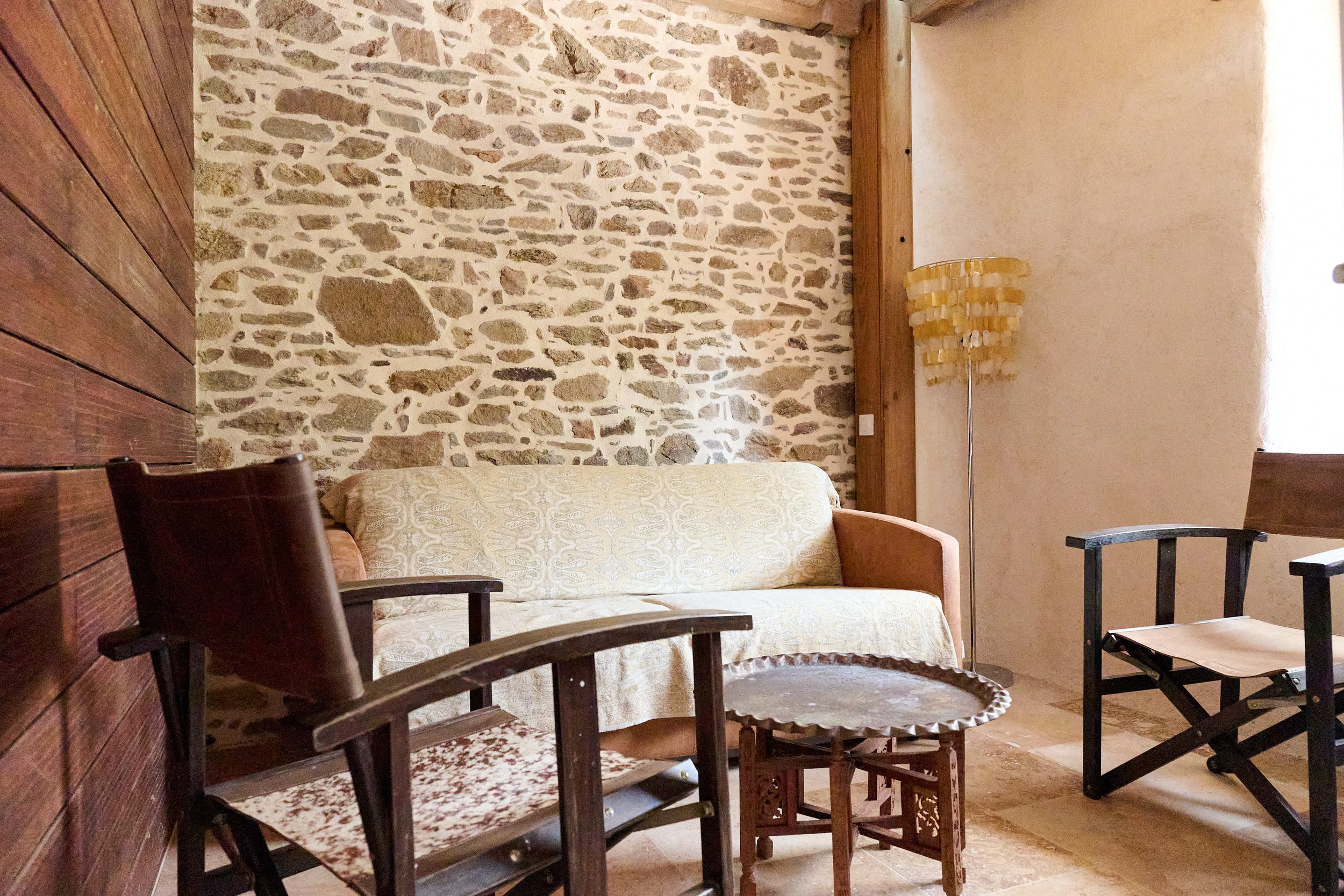 Interior of a room with a stone wall, a wooden ceiling, and a beige sofa. There are chairs around a round wooden table, a standing lamp with a yellow shade, and a window on the right side.