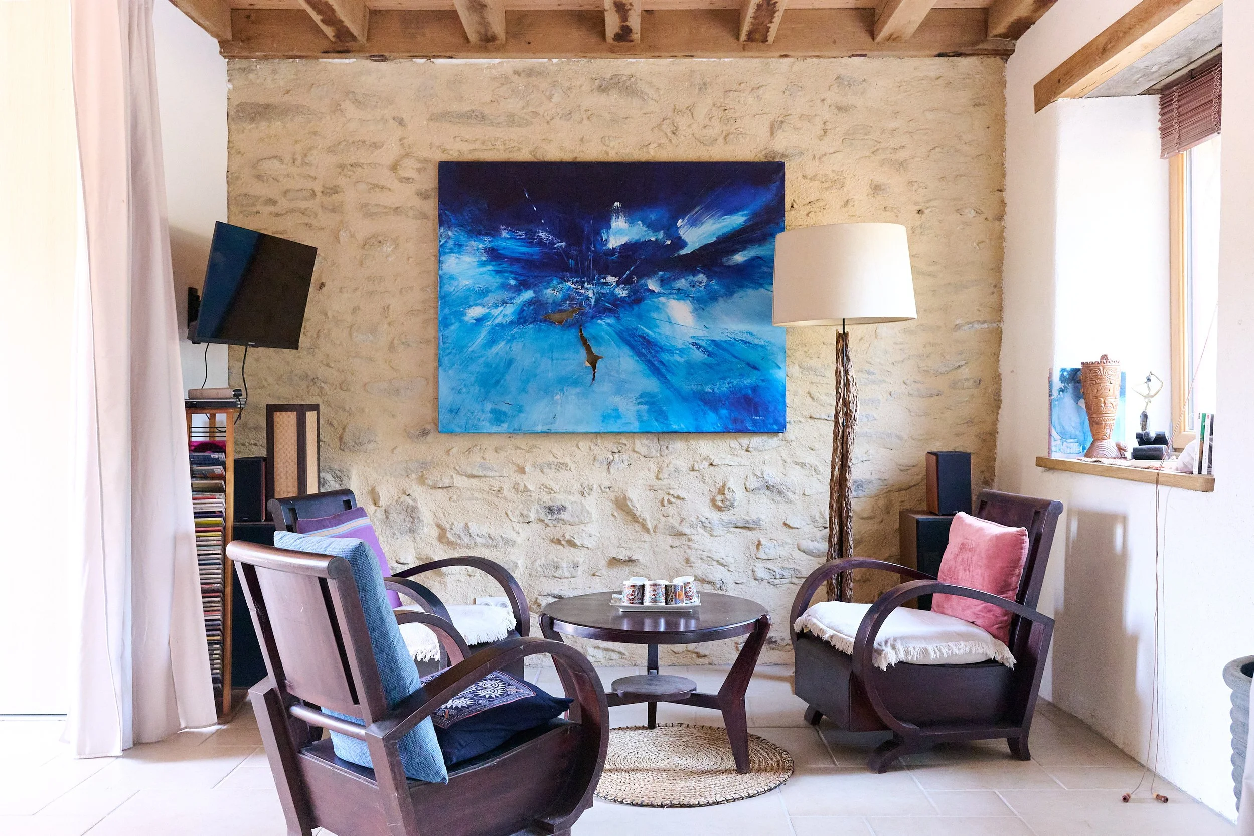 Interior of a living room with a stone wall, wooden ceiling beams, a large abstract blue painting, a flat-screen TV, a floor lamp, and various chairs and decorations.