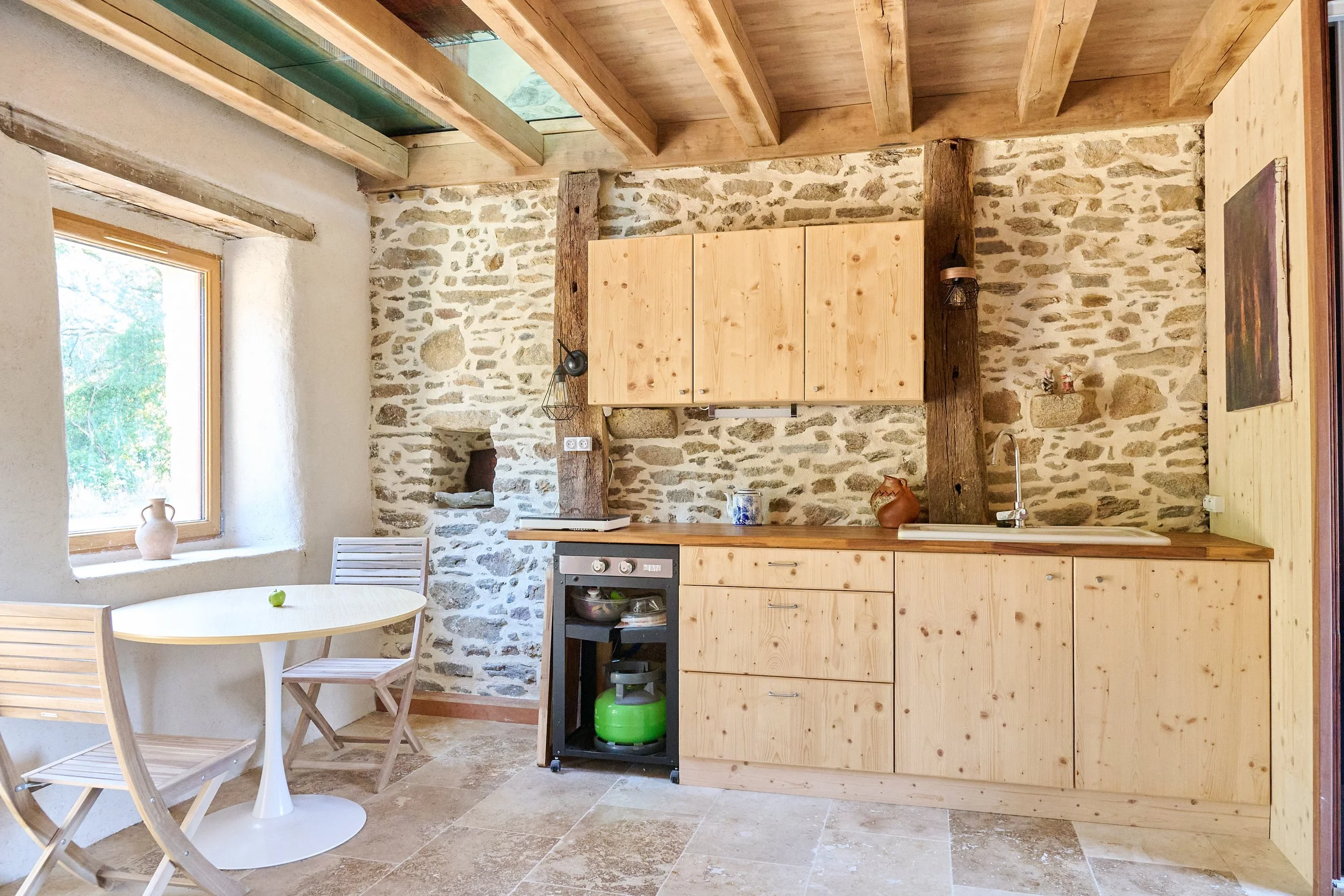 A rustic kitchen with stone walls, wooden beams, a small round dining table with wooden chairs, a window with a ceramic vase, and kitchen cabinets with a stove and green propane tank underneath.