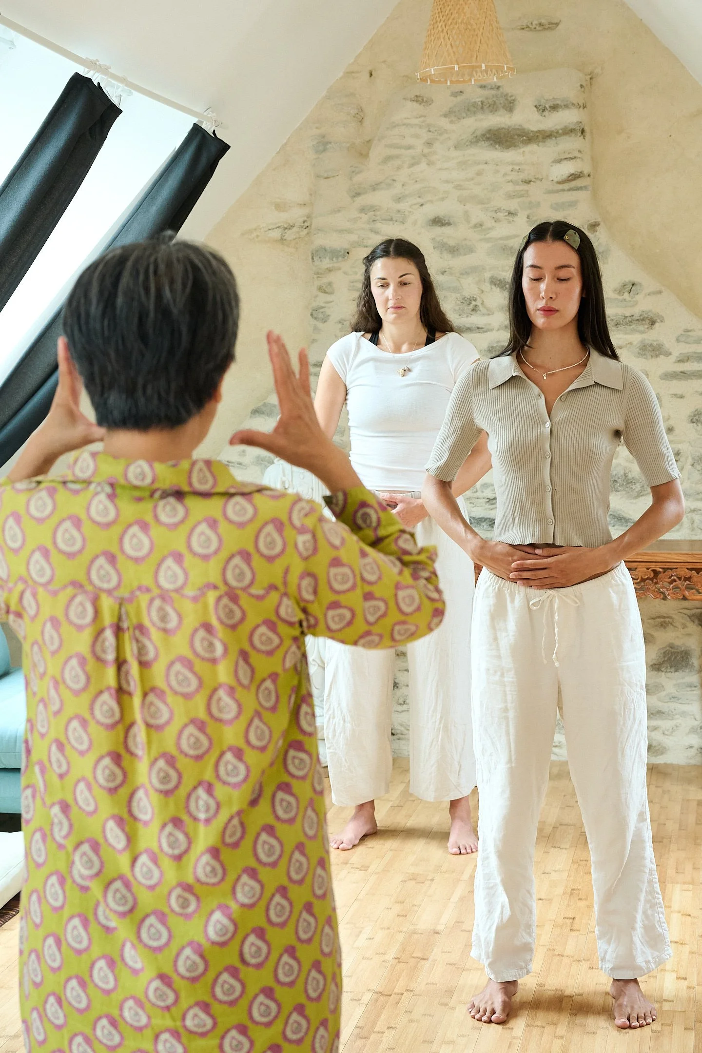 Three women participating in a yoga or meditation class in a room with stone wall and wooden flooring. One woman is leading, others are following her pose.