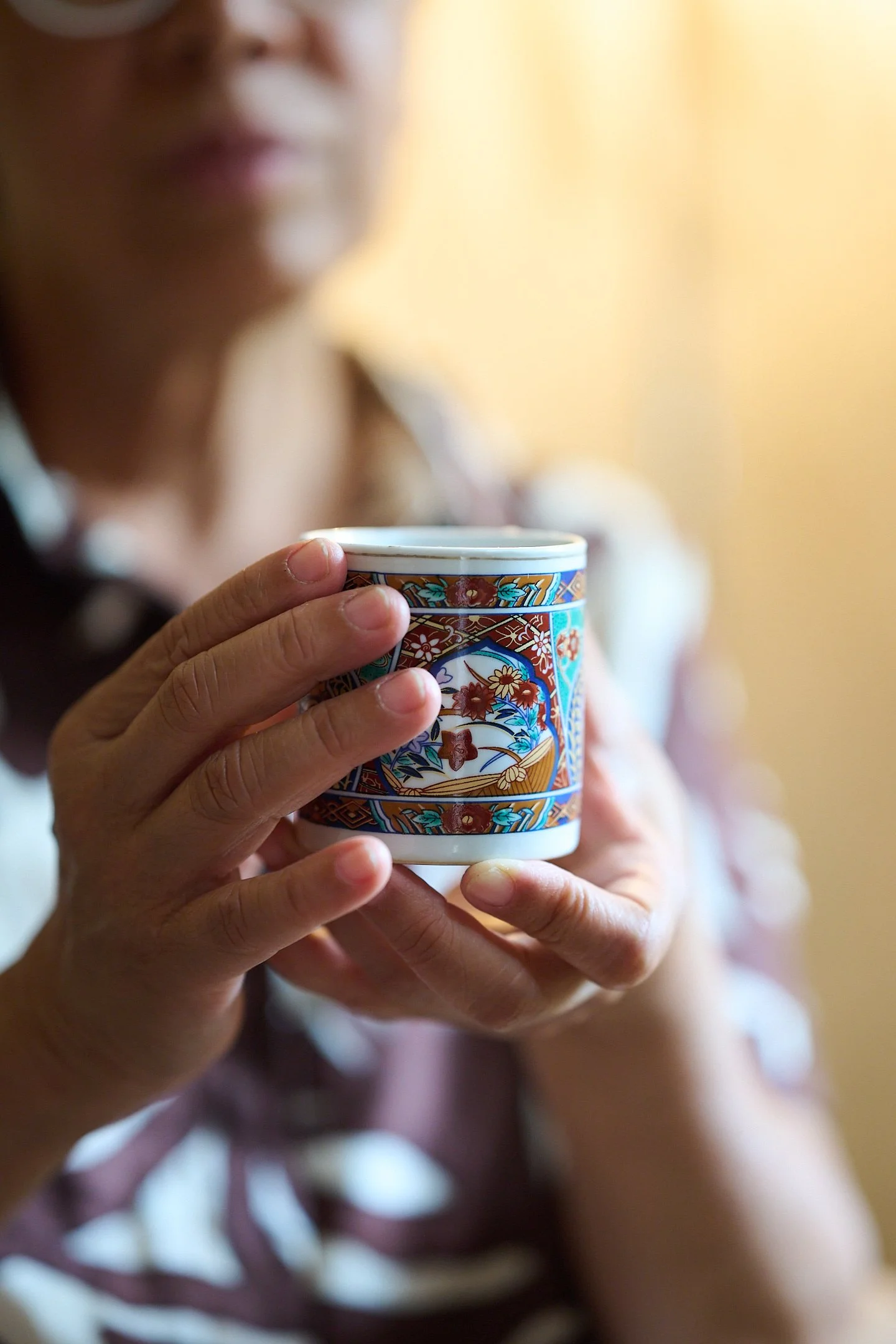 A person holding a colorful ceramic mug with intricate patterns, with a blurred face in the background.