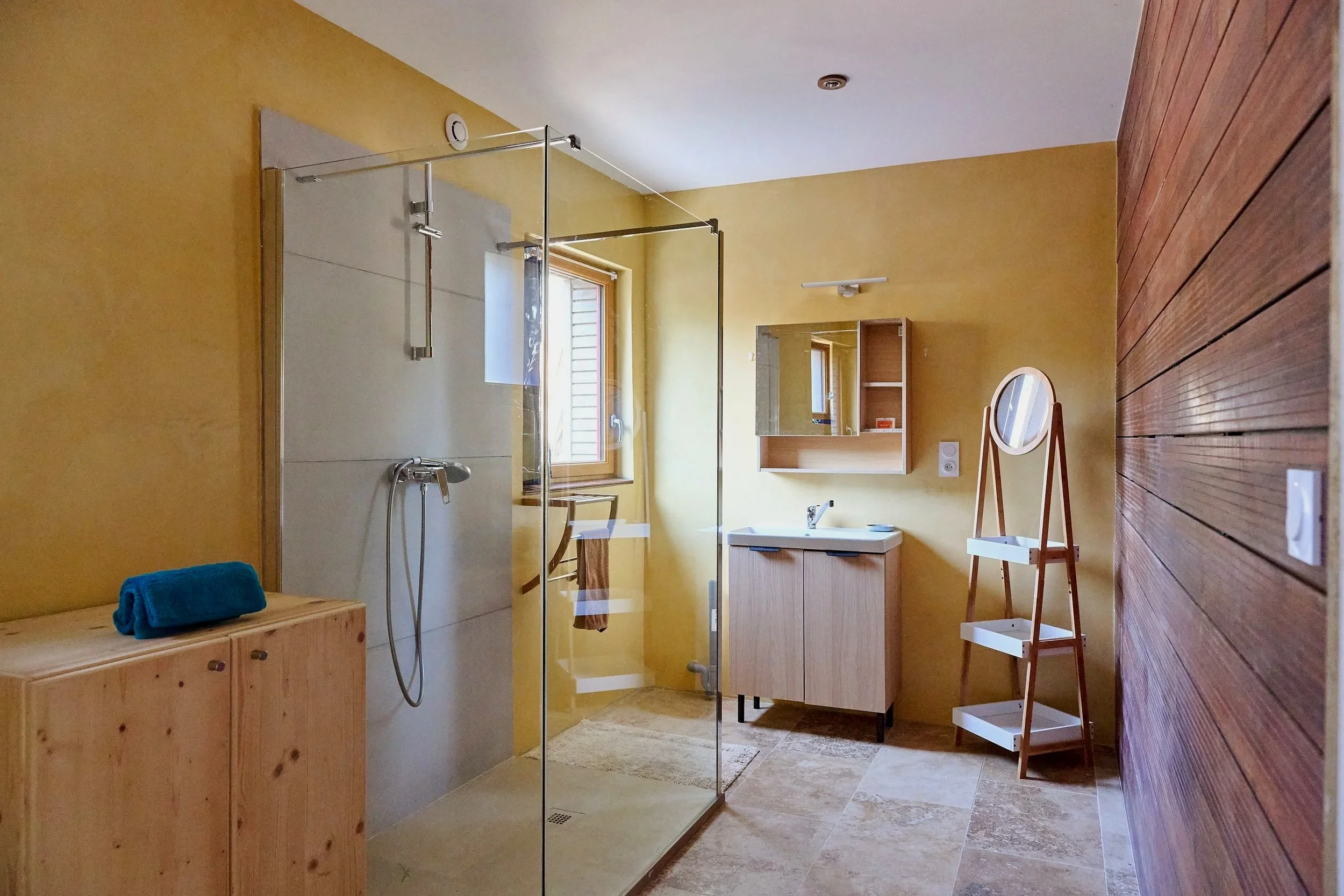 A bathroom with yellow walls, a tiled floor, and a wooden ceiling. It features a glass-enclosed shower with a silver showerhead, a window with natural light, a wooden cabinet, and a small mirror. There is also a towel on a wooden bench and a white ladder shelf.
