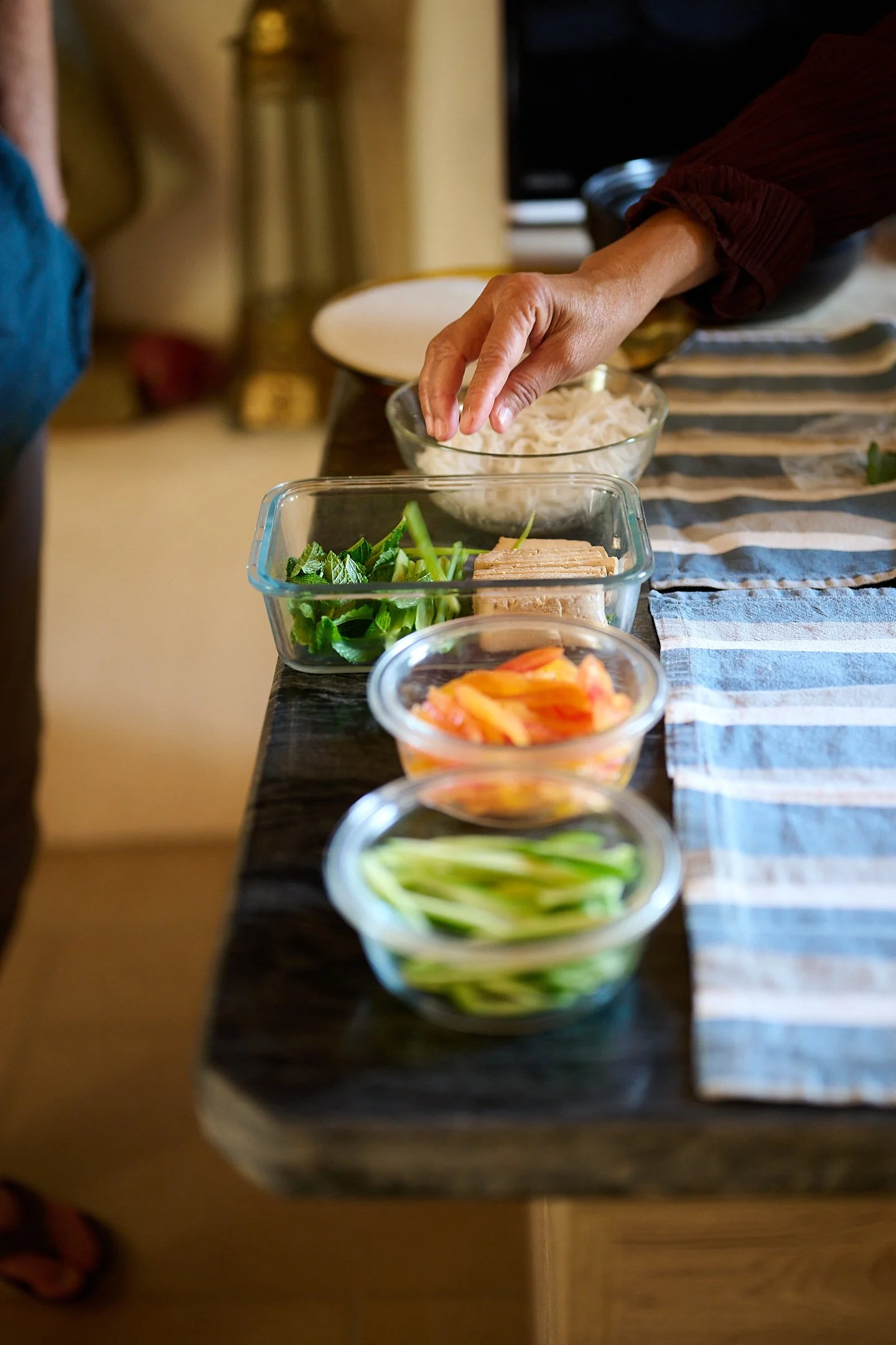 A person preparing ingredients for a meal with bowls containing vegetables, noodles, and other food items on a table.