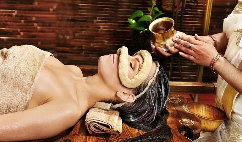 A woman receiving a facial treatment at a spa, with a towel wrapped around her torso, lying on a wooden massage table, with her eyes closed and a towel rolled under her head. A staff member is holding a bowl with a dark liquid, possibly part of the treatment, with plants and wooden decor in the background.