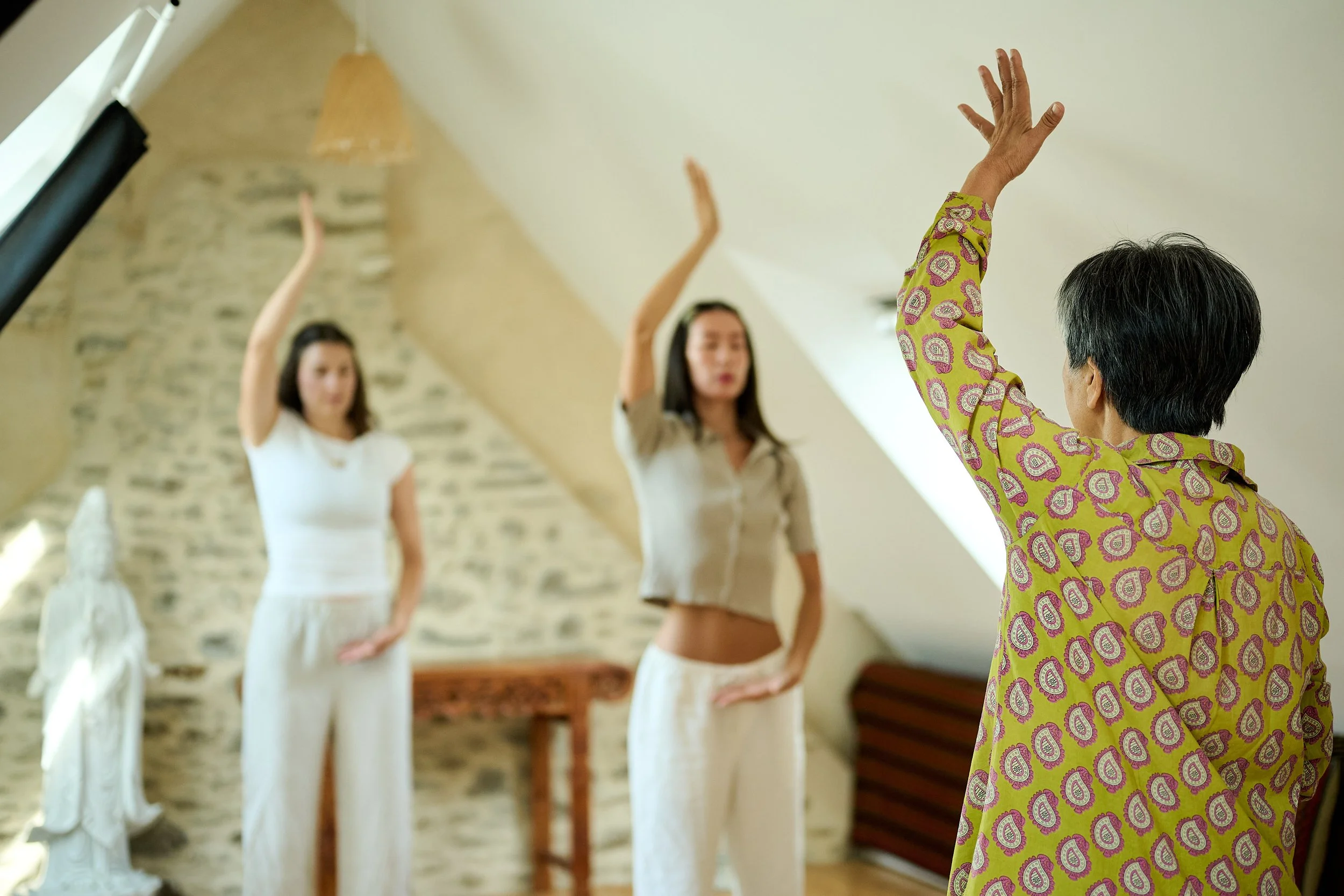 A person in a colorful paisley shirt raising their hand in front of three women standing with hands raised in a room with stone walls and a sloped ceiling.