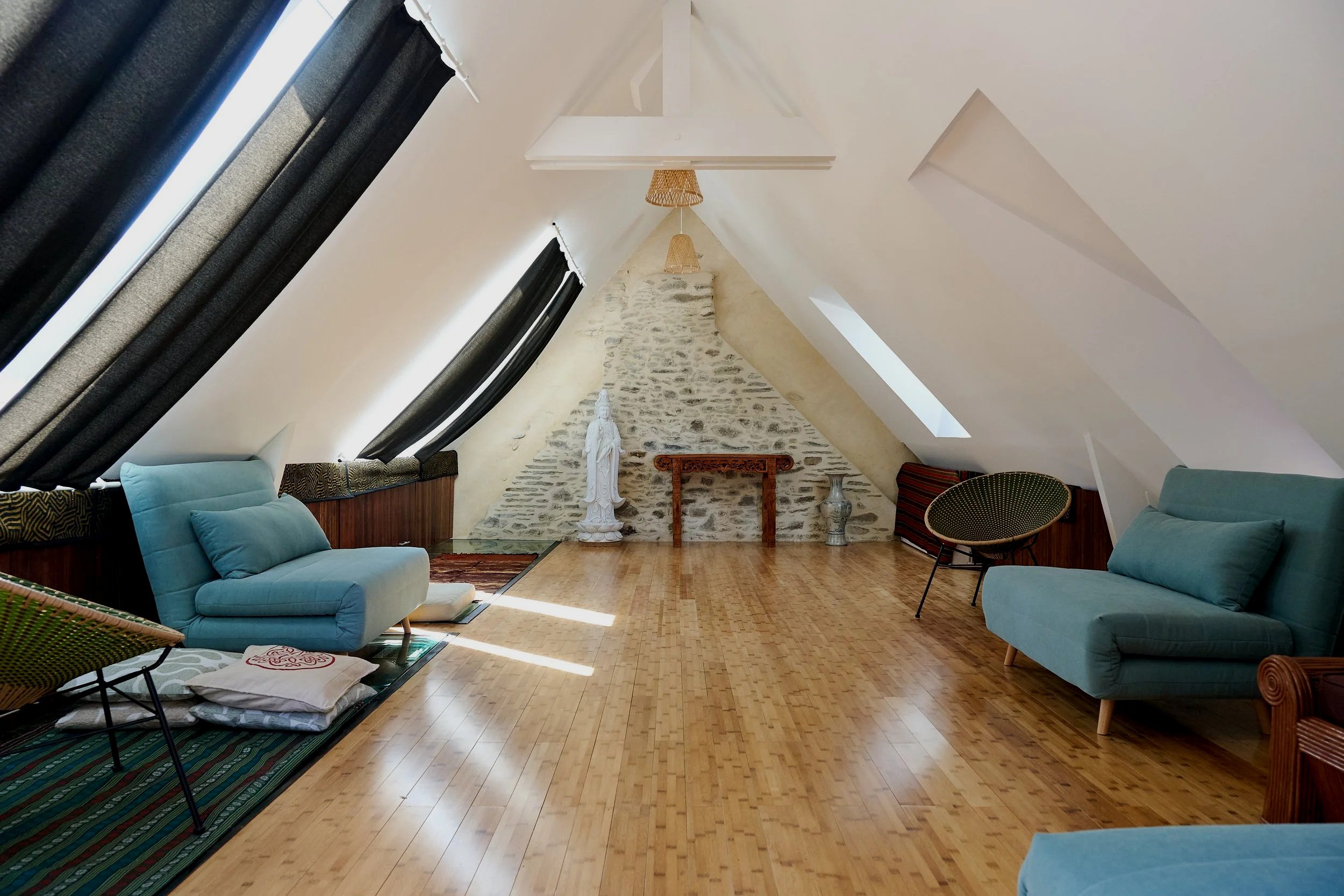 A cozy attic living room with sloped white ceilings, natural light from skylights, and a stone accent wall. The room features blue upholstered furniture, a wooden floor, and various decorative items including a statue, vases, and a small dining table.