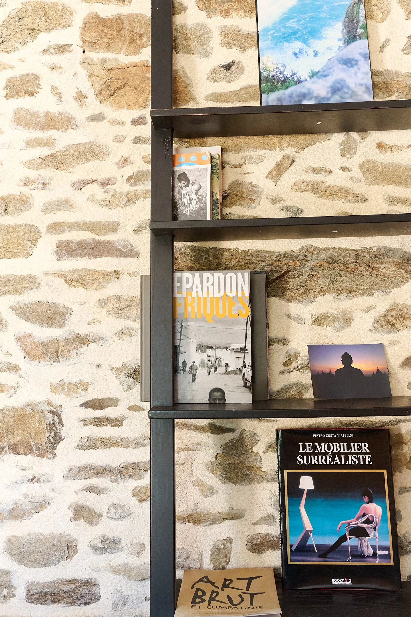 Books and artwork displayed on a black metal shelf against a stone wall, including a magazine titled 'LE MOBILIER SURREALISTE' with a photograph of a woman sitting next to a lamp at sunset.