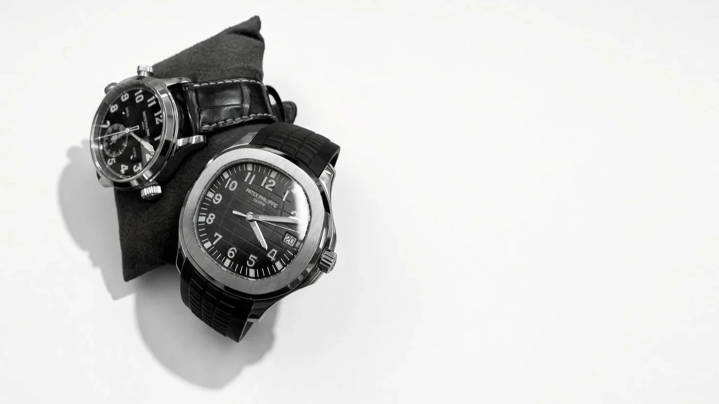 Two luxury wristwatches placed on a black cushion against a white background. One has a black leather strap and the other has a black rubber strap.