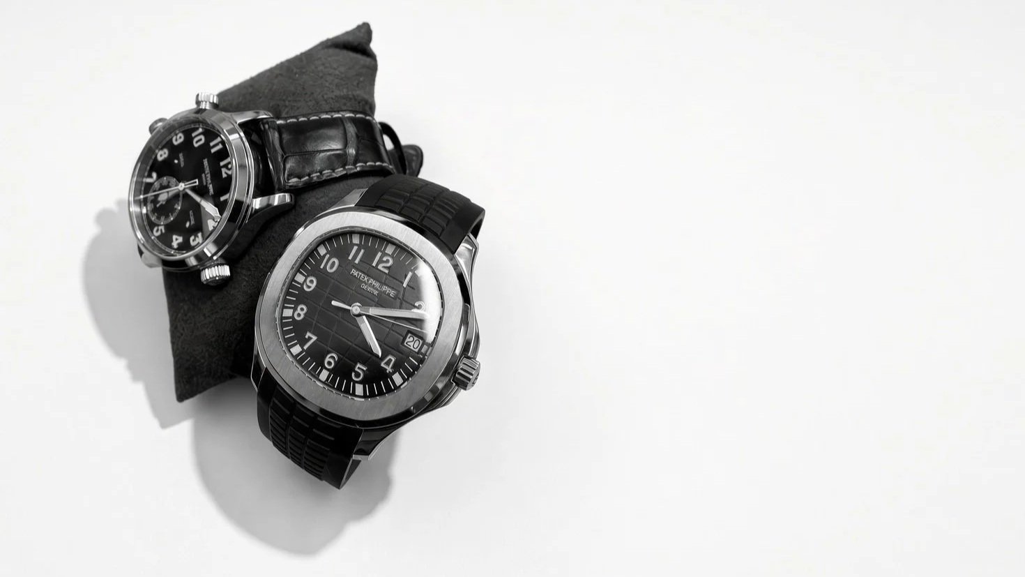 Two luxury wristwatches placed on a black cushion against a white background. One has a black leather strap and the other has a black rubber strap.