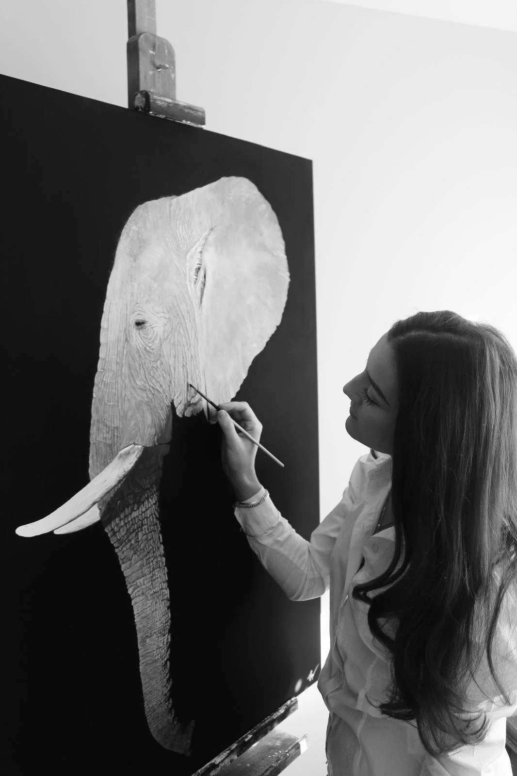 A woman painting an elephant on a black canvas, seen from the side, with the woman focusing on details of the elephant's face.