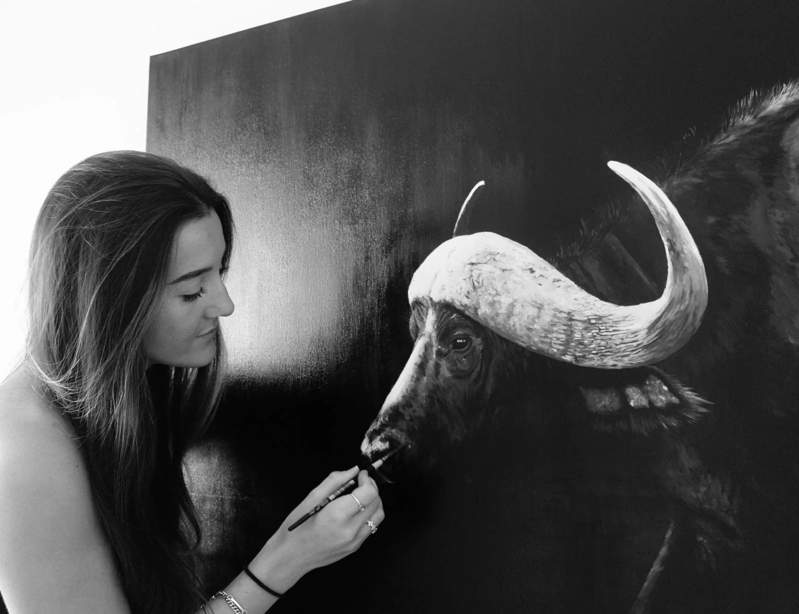 A woman with long hair painting a realistic black and white portrait of a buffalo on a dark wall.