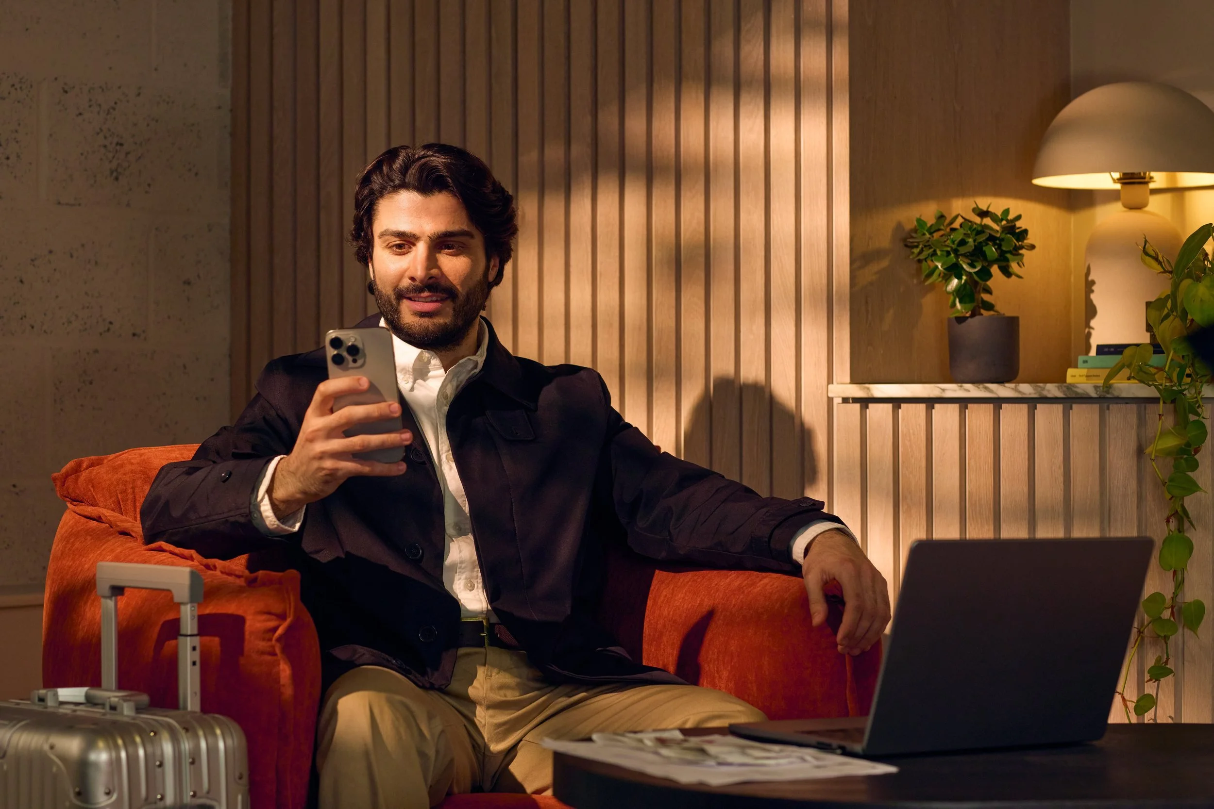 A man with dark hair and beard sitting on an orange couch, looking at his phone with a smile. He is dressed in a dark blazer, white shirt, and khaki pants. The room has wooden paneling, a table with a laptop, and a luggage case nearby. A lamp, potted