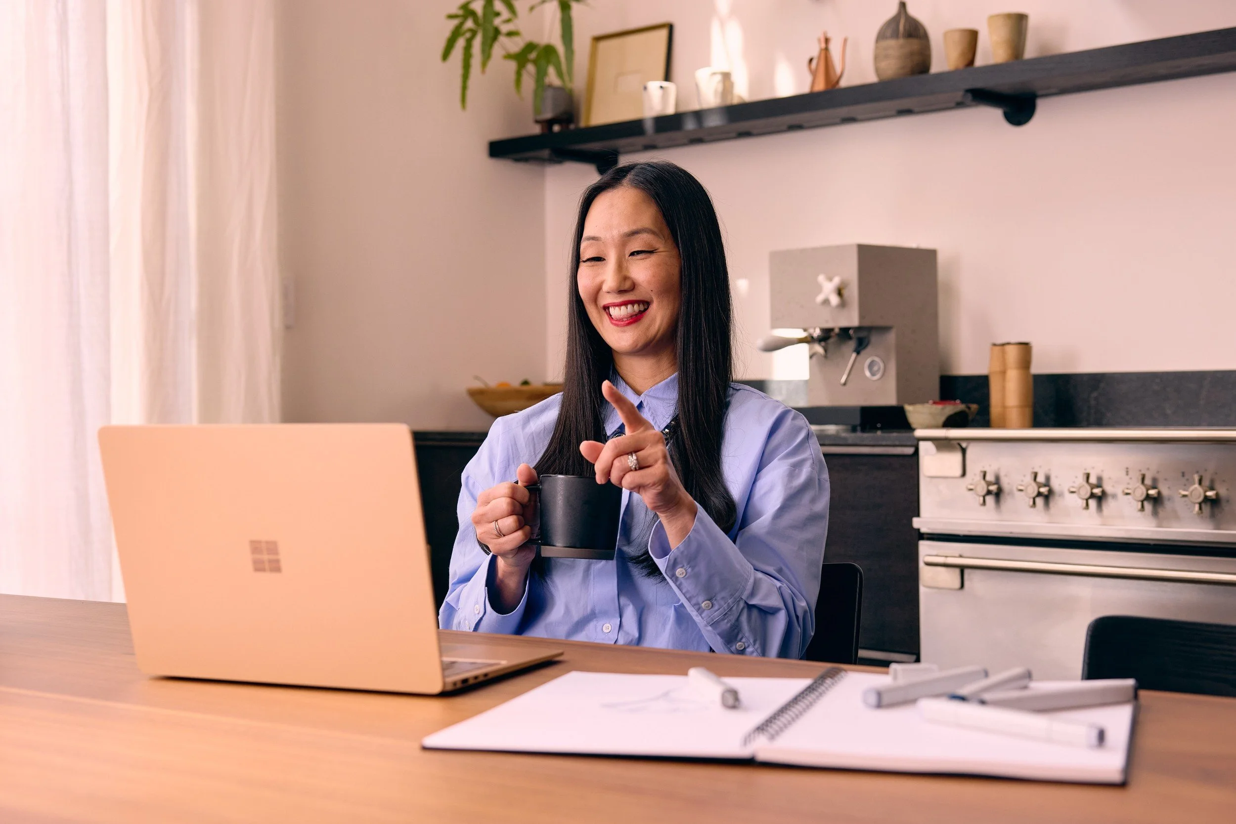 A woman sitting at a desk, smiling, holding a coffee mug, with a laptop and notebooks in front of her, in a cozy kitchen or dining area.