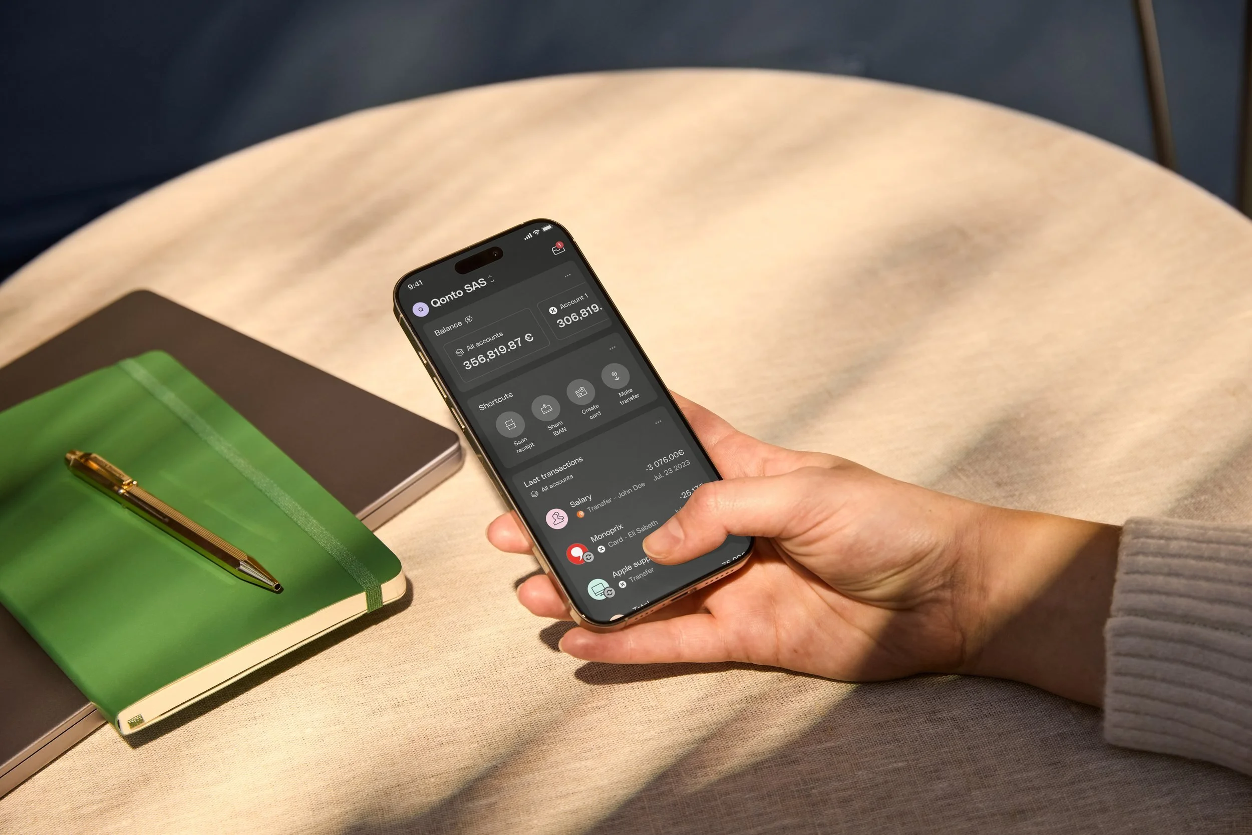 Person holding a smartphone showing a bank account balance, with a green notebook and pen on the table.