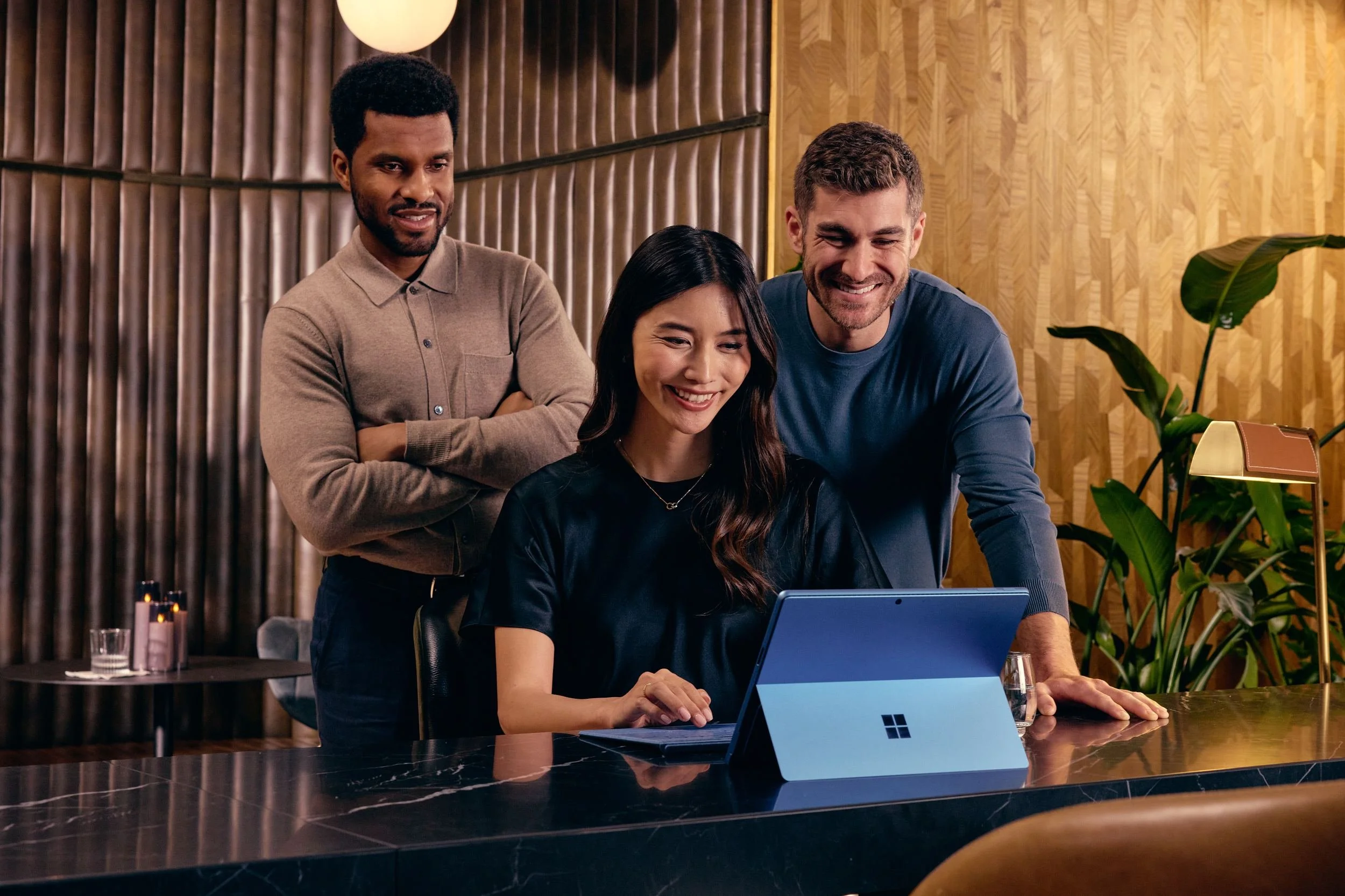 Three people in a modern office smiling at a laptop, with one woman seated and two men standing behind her, surrounded by wooden and green decor.
