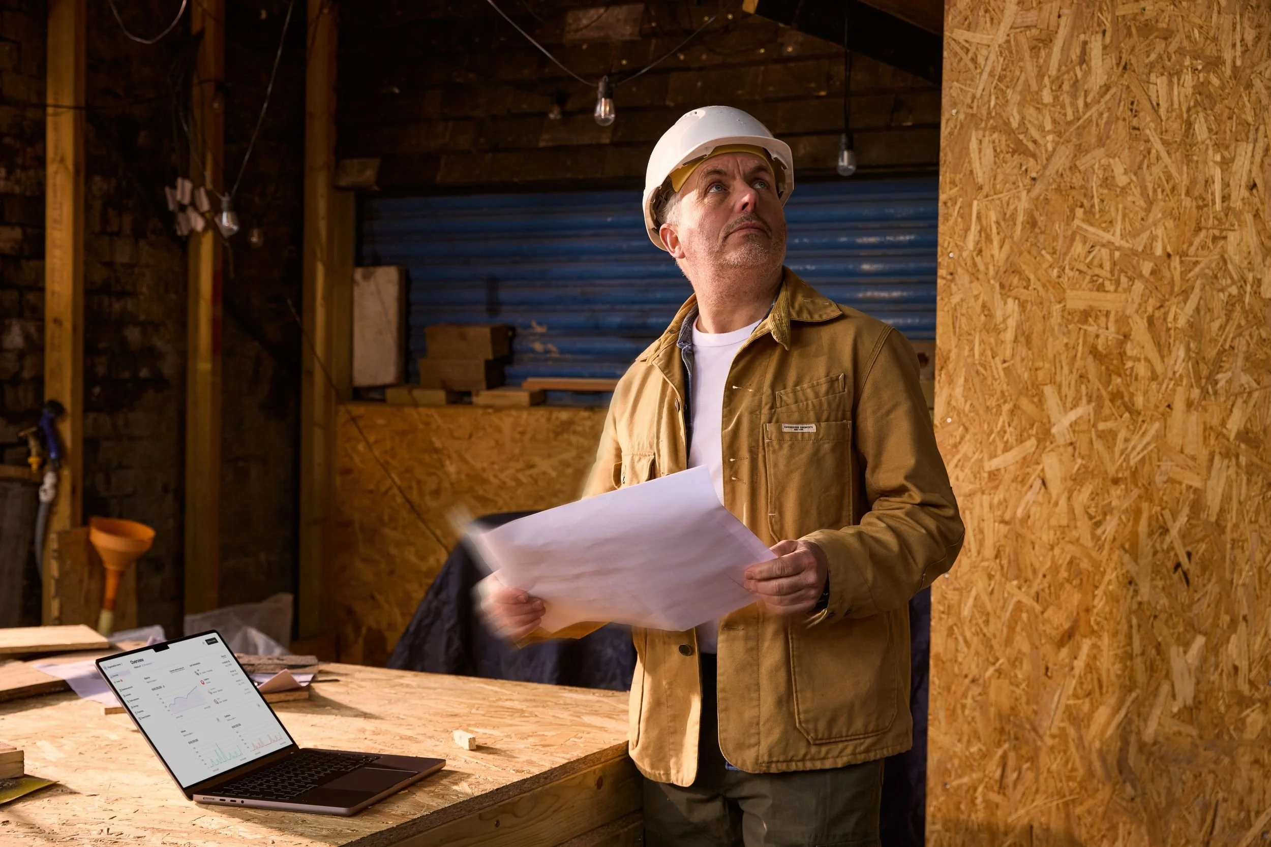 A man in a construction helmet and jacket holding papers inside a workshop or construction site with wooden walls and a table with a laptop displaying charts.