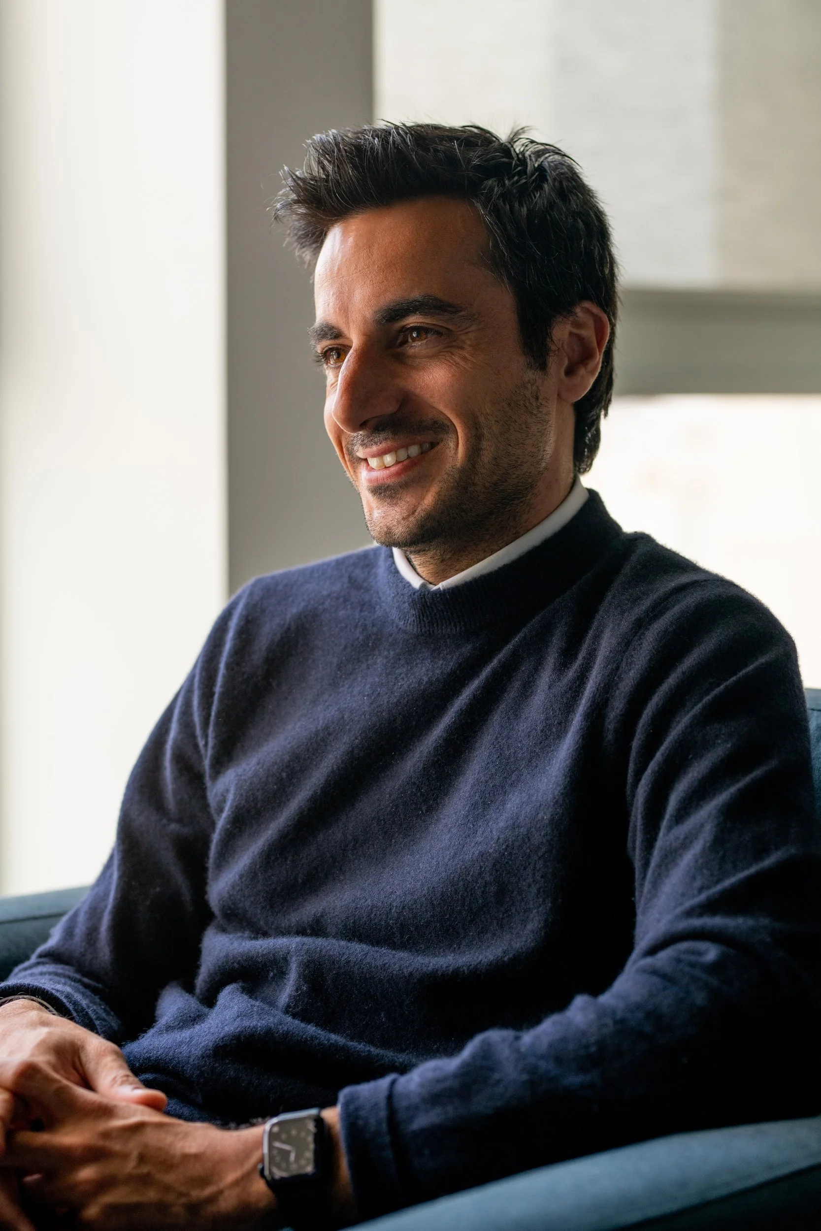 A man with dark hair and a beard smiling while sitting indoors near a window, wearing a dark sweater and a smartwatch on his left wrist.