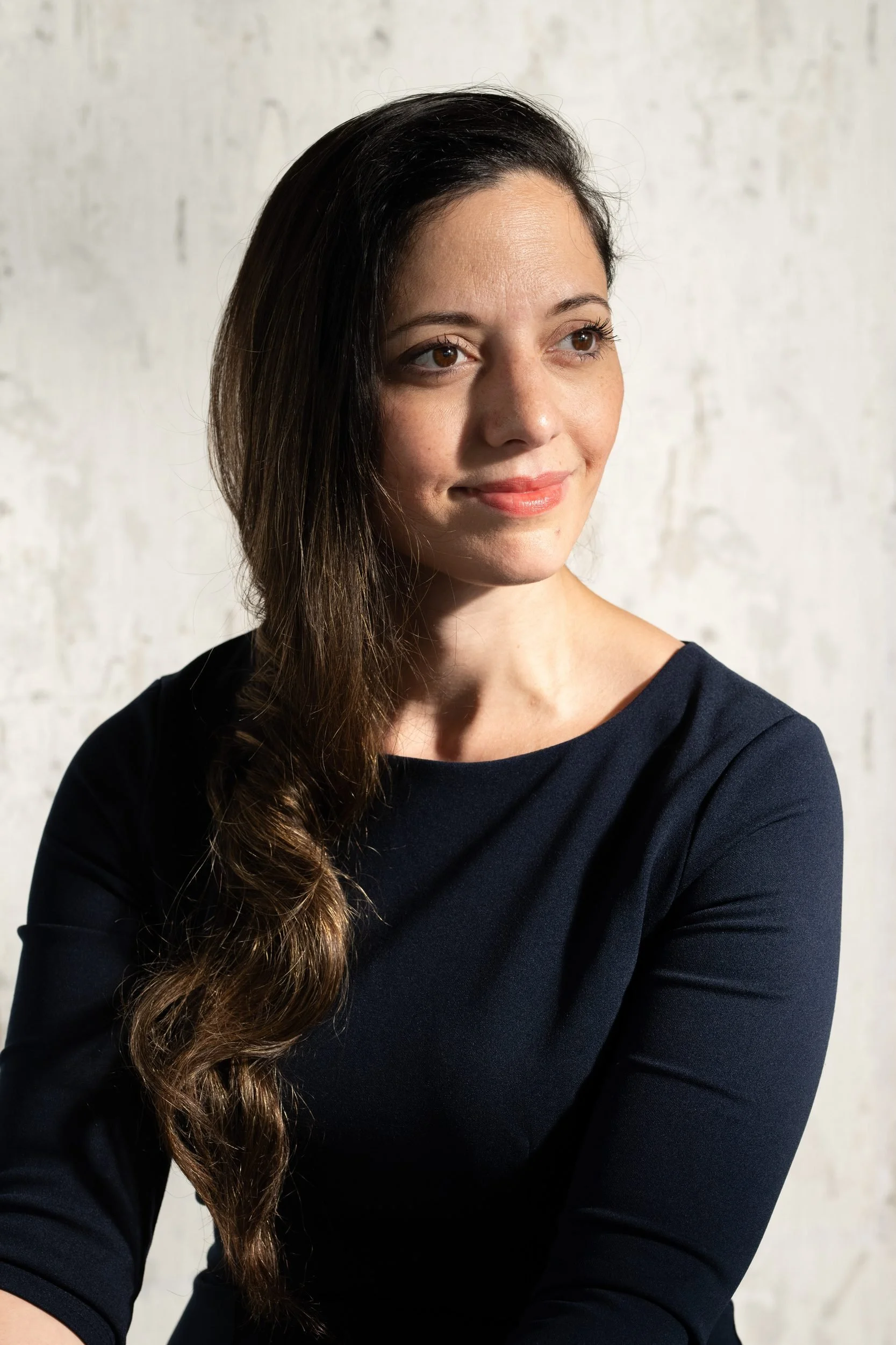 A woman with long dark hair and brown eyes, wearing a black top, looking to the right against a light textured background.
