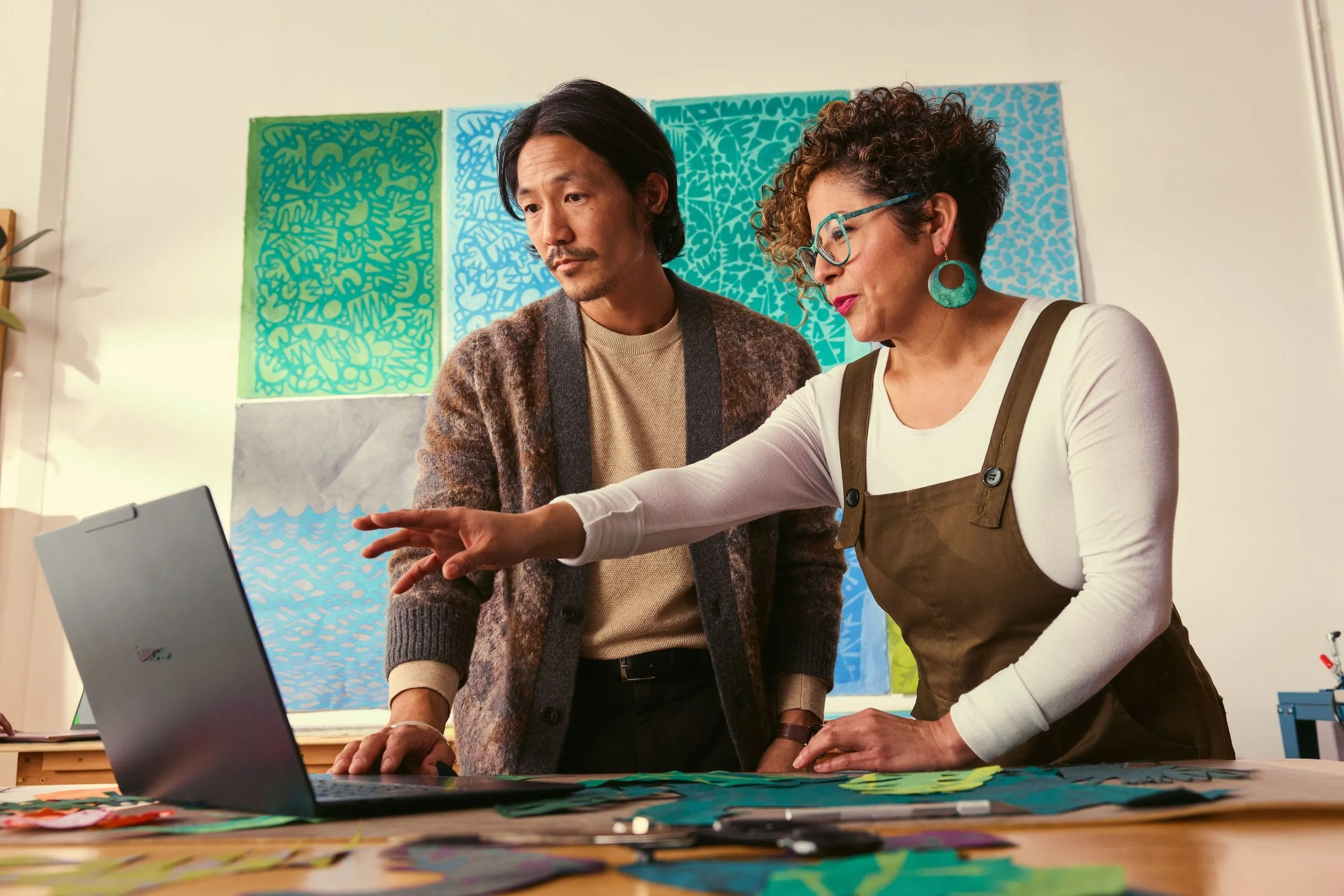 Two individuals, a man and a woman, are working together on a laptop in an art studio. The woman is pointing at the laptop screen, and both are focused on it. The background includes colorful, patterned artwork.