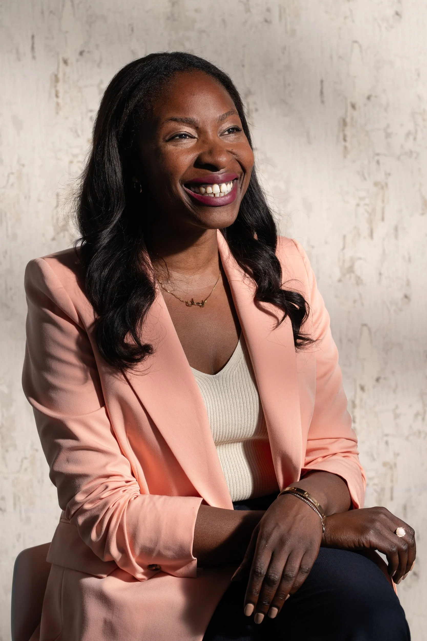 A smiling woman with dark, wavy hair, wearing a light pink blazer over a beige top, sitting and resting her hands on her lap, against a textured light-colored wall background.