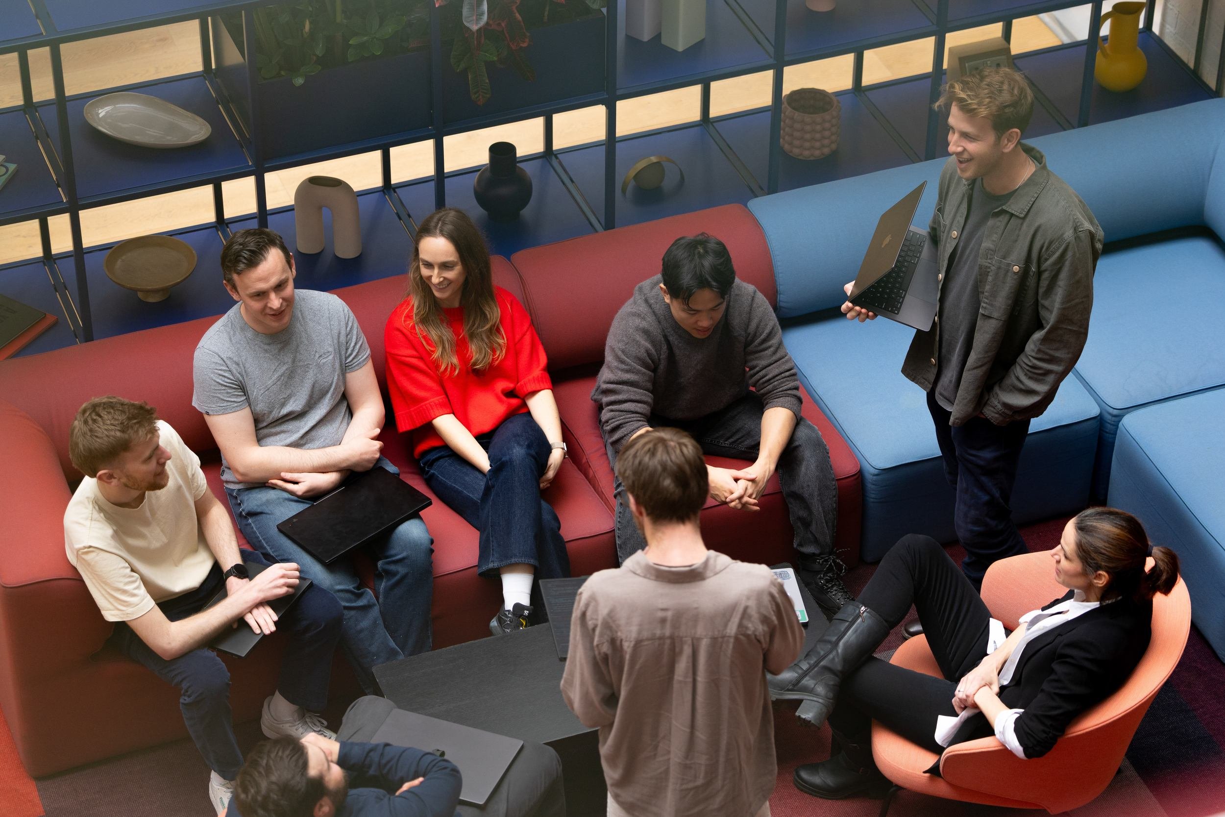 Group of young adults sitting on a red sofa and chairs in a modern office or coworking space, engaged in a discussion or meeting, with one young man standing, holding a laptop and smiling.