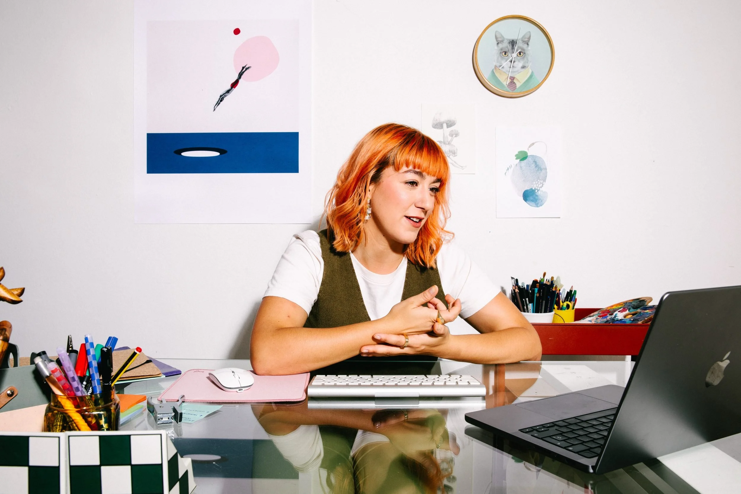 A young woman with orange hair sitting at a desk in front of a laptop, surrounded by art supplies and stationery, in a well-lit room with artwork and a clock with a cat picture on the wall behind her.