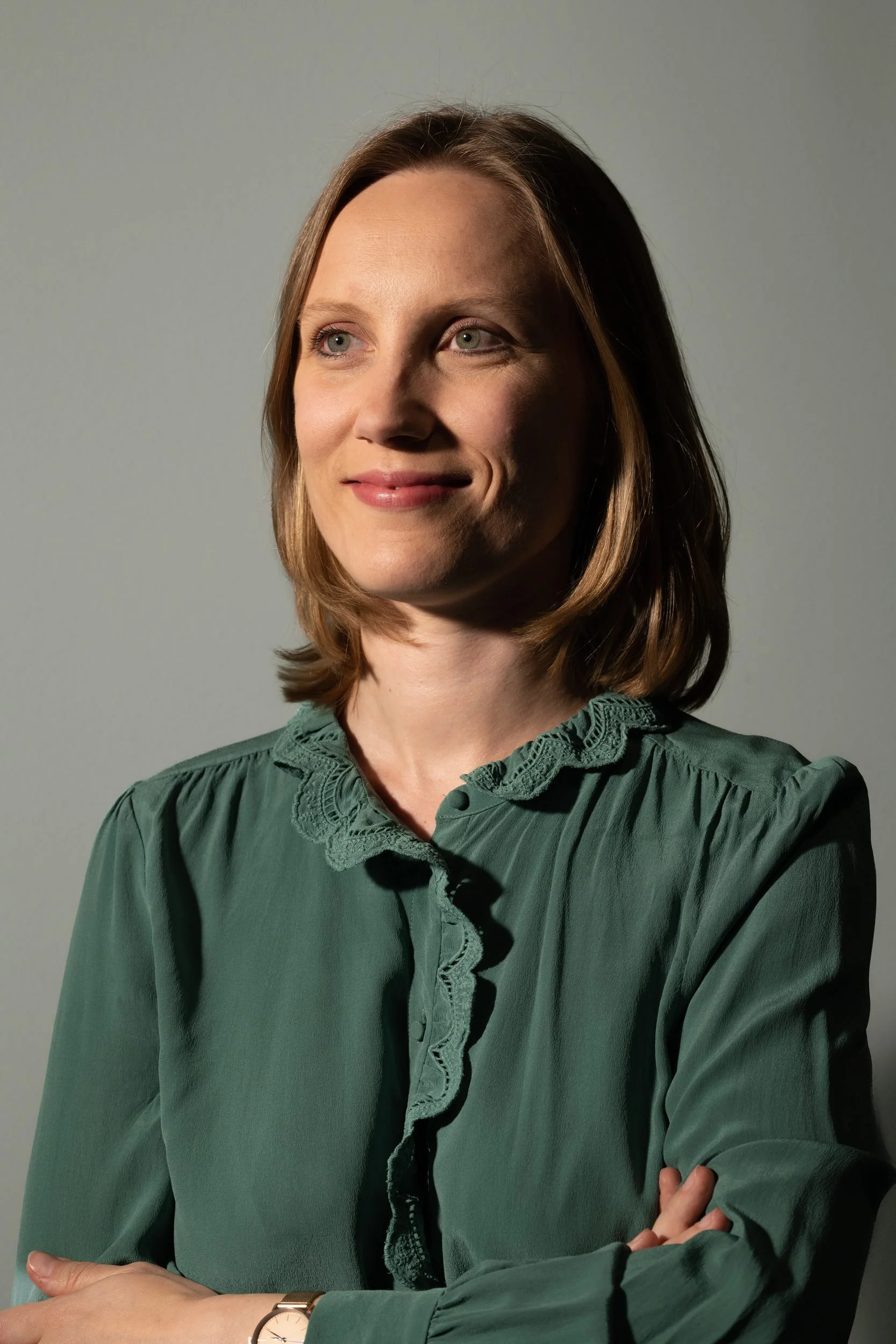 Woman with shoulder-length brown hair wearing a green blouse with lace details, standing against a gray background with arms crossed.