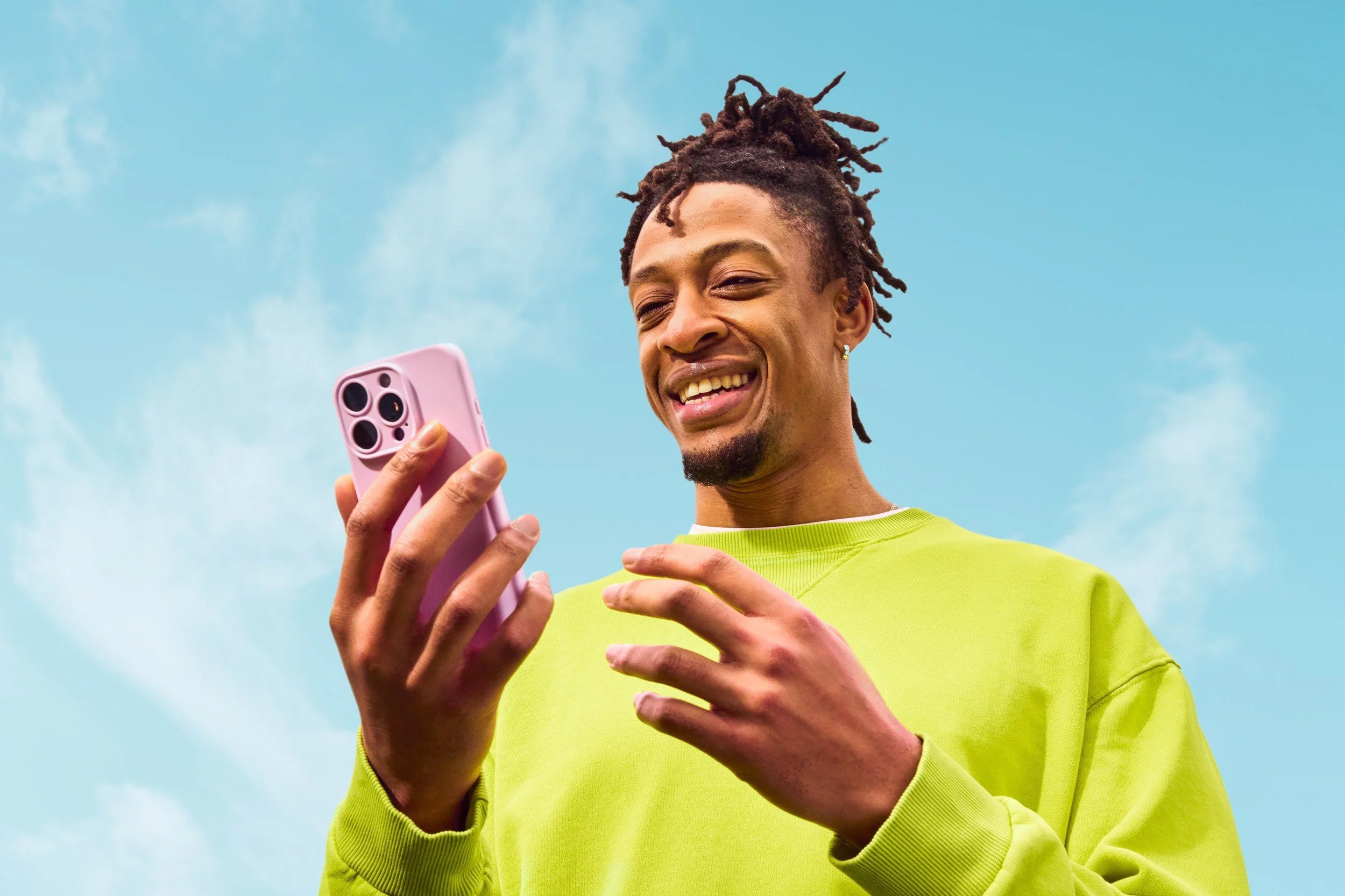 A young man with dreadlocks, wearing a bright yellow-green sweatshirt, smiling while looking at his pink smartphone against a blue sky with scattered white clouds.