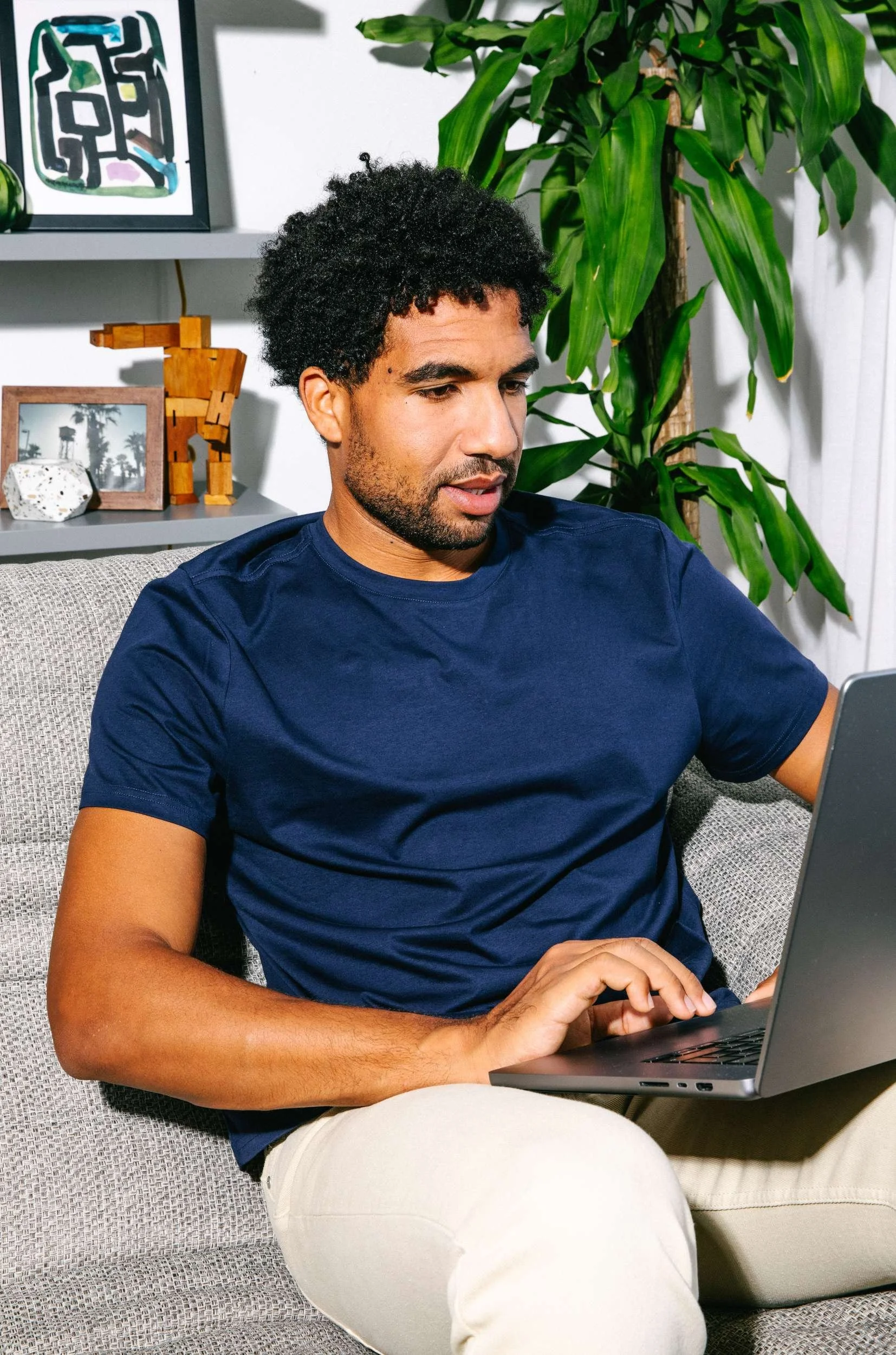 A man with curly black hair and a beard sitting on a gray patterned sofa, wearing a navy blue t-shirt and white pants, working on a laptop. There are green plants and framed pictures on shelves behind him.