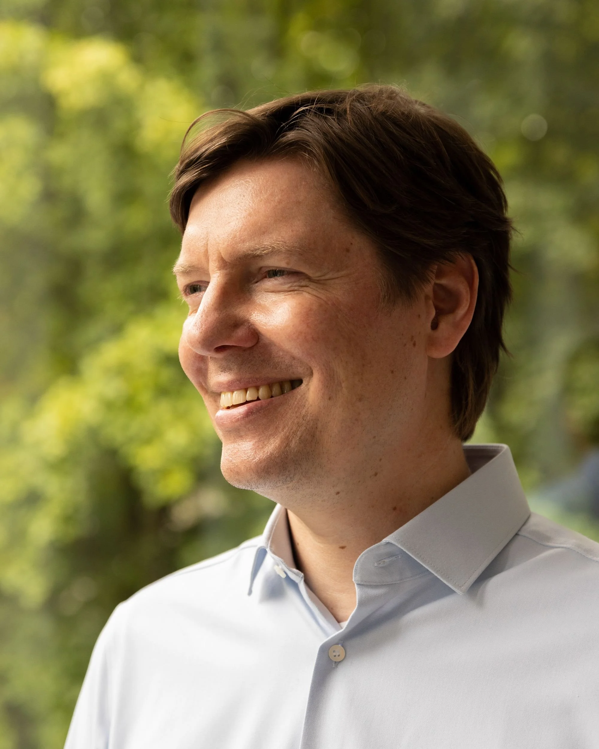 Close-up of a smiling man wearing a light blue collared shirt, outdoors with a blurred green background.