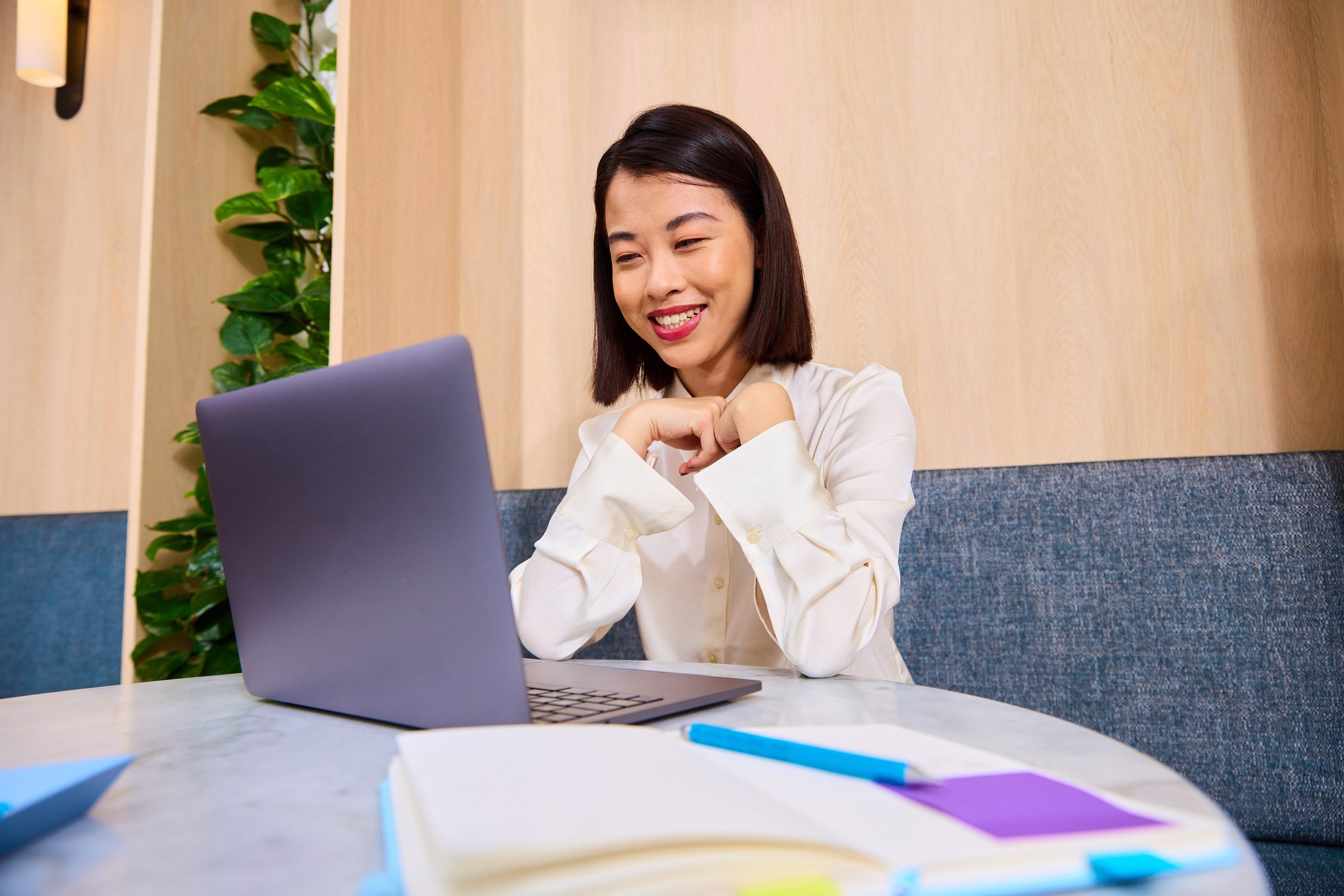 A young woman with dark hair sitting at a table in a modern office, smiling and looking at her laptop, with notebooks and pens on the table.