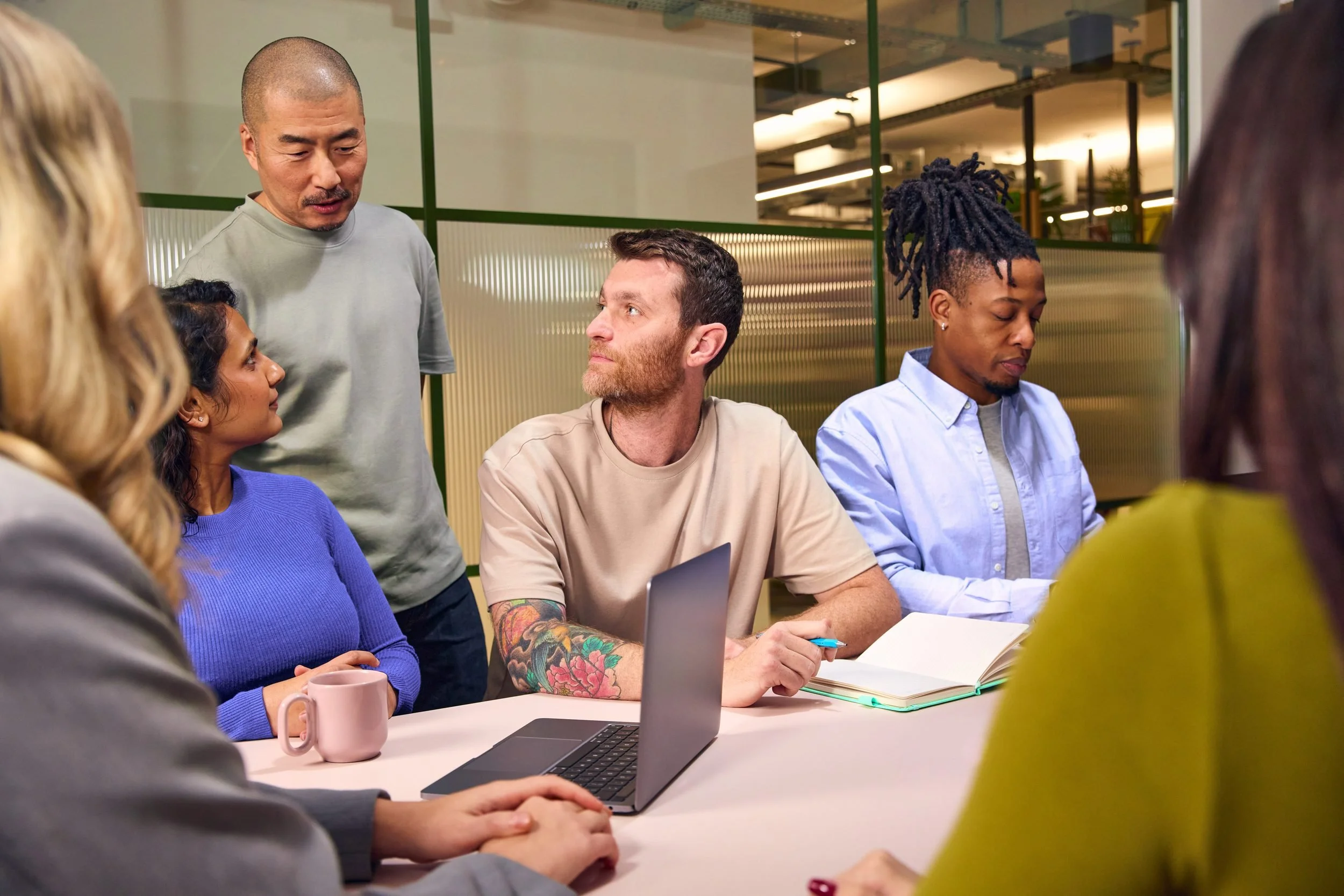 Group of diverse people sitting around a table in a meeting or discussion, with laptops, notebooks, and a mug visible, in a modern office environment.