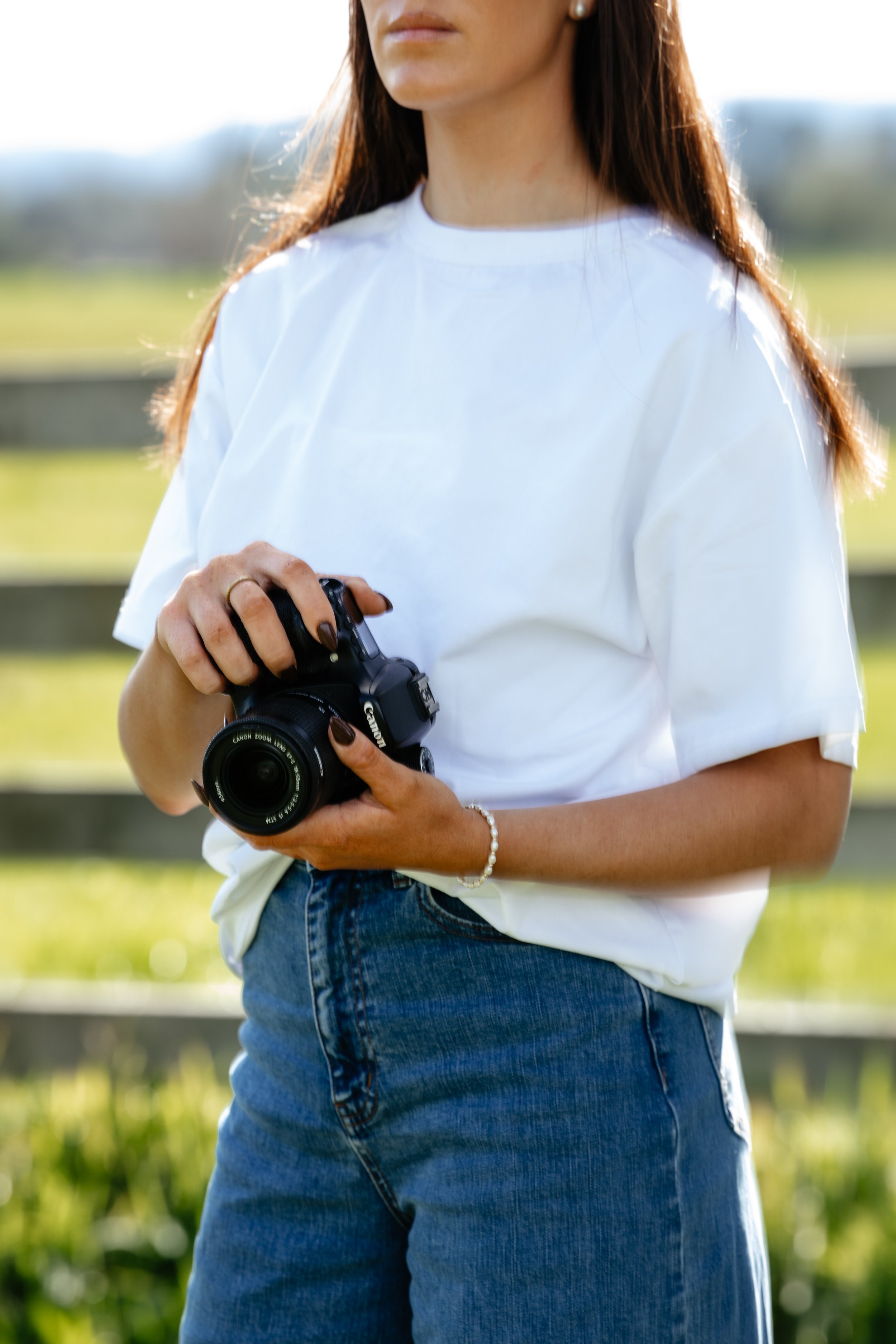 A woman wearing a white t-shirt and blue jeans holding a black camera outdoors with a blurred green field background.
