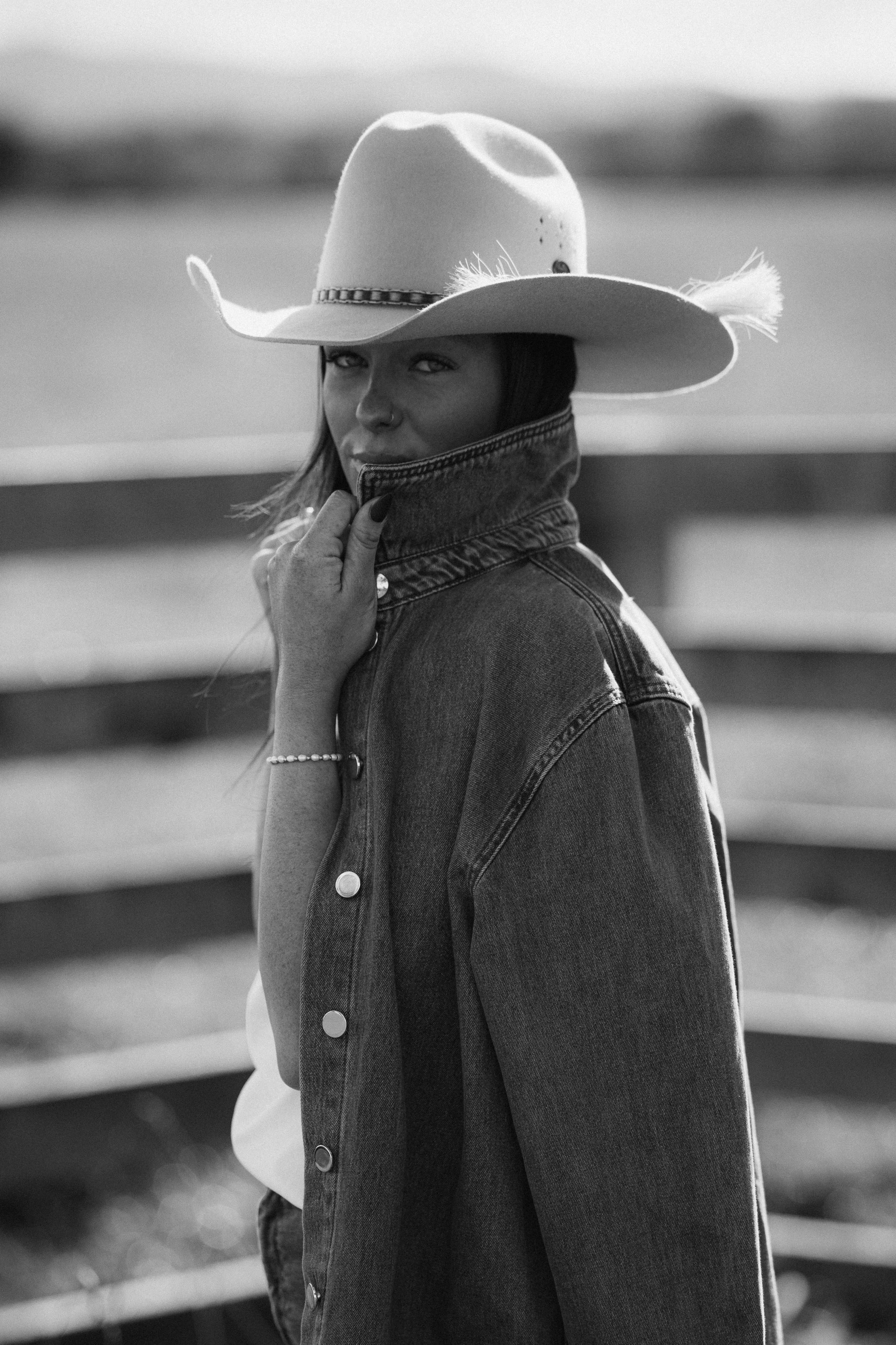 Black and white photo of a woman wearing a wide-brimmed cowboy hat and denim jacket, looking over her shoulder with a serious expression, outdoors with blurred background.