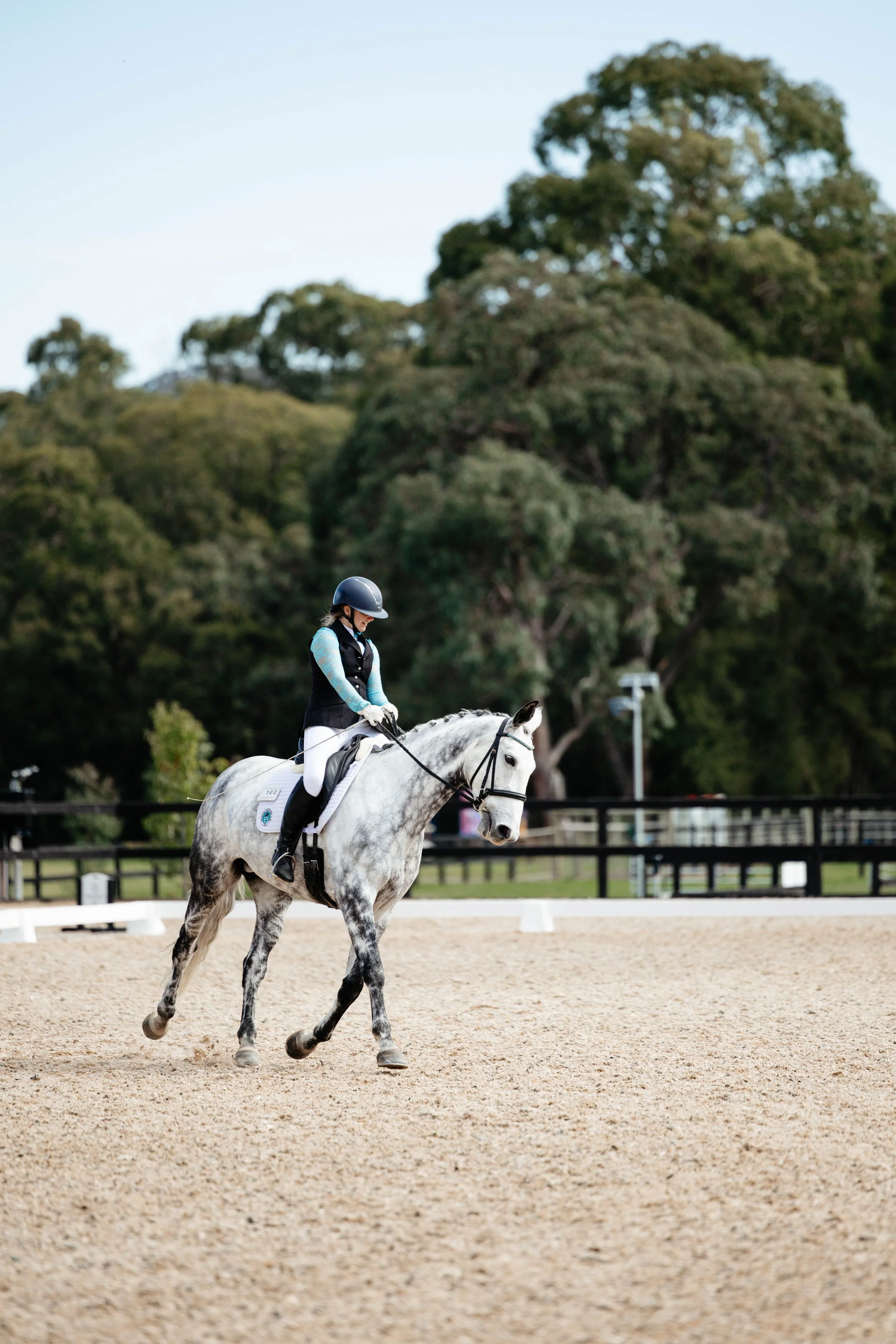A woman riding a gray horse in an outdoor riding arena with trees in the background.