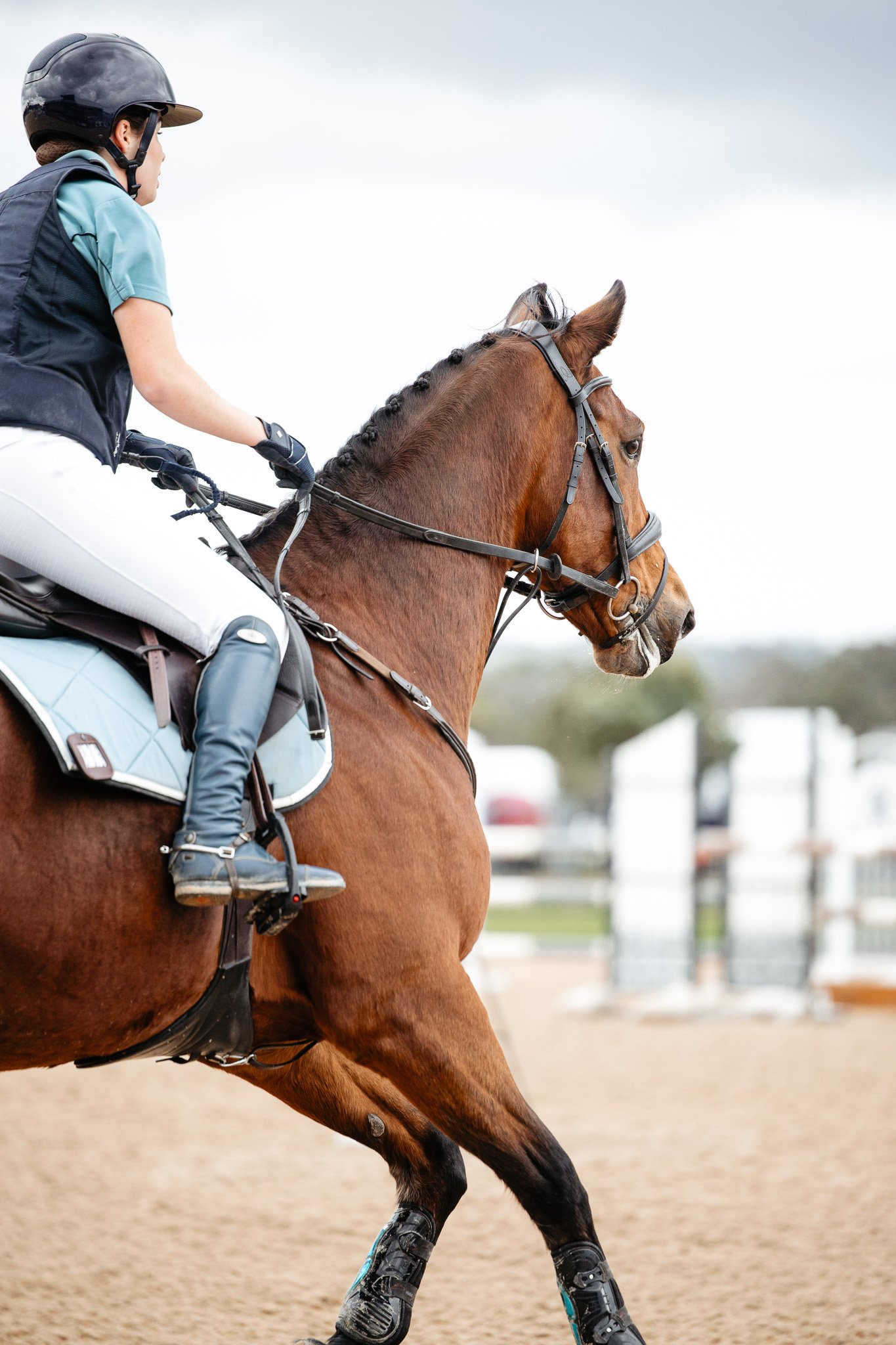 A person riding a brown horse at a riding arena, wearing a helmet, light blue shirt, and riding gear.