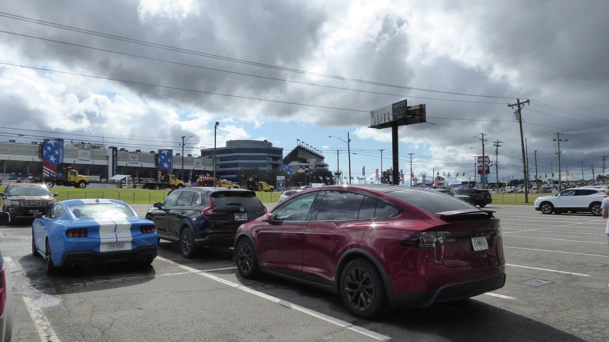 Our red Tesla Model X parked beside a blue Ford Mustang at Charlotte Motor Speedway, with the grandstands and race-weekend activity in the background.