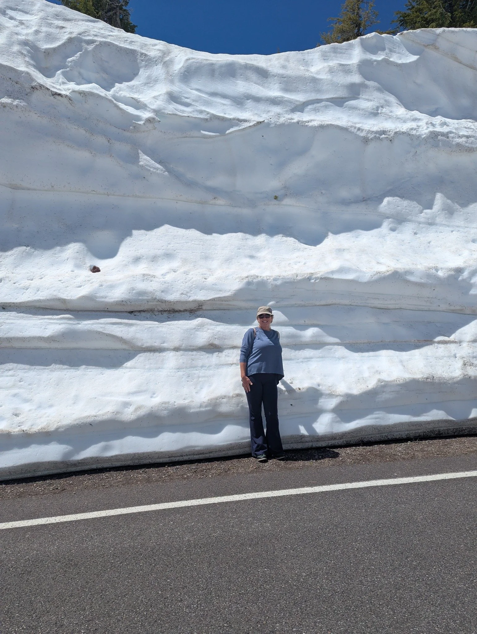 Gina standing beside an enormous plowed snowbank along a clear road at Crater Lake National Park in late May, showing the remarkable height of the remaining snow.