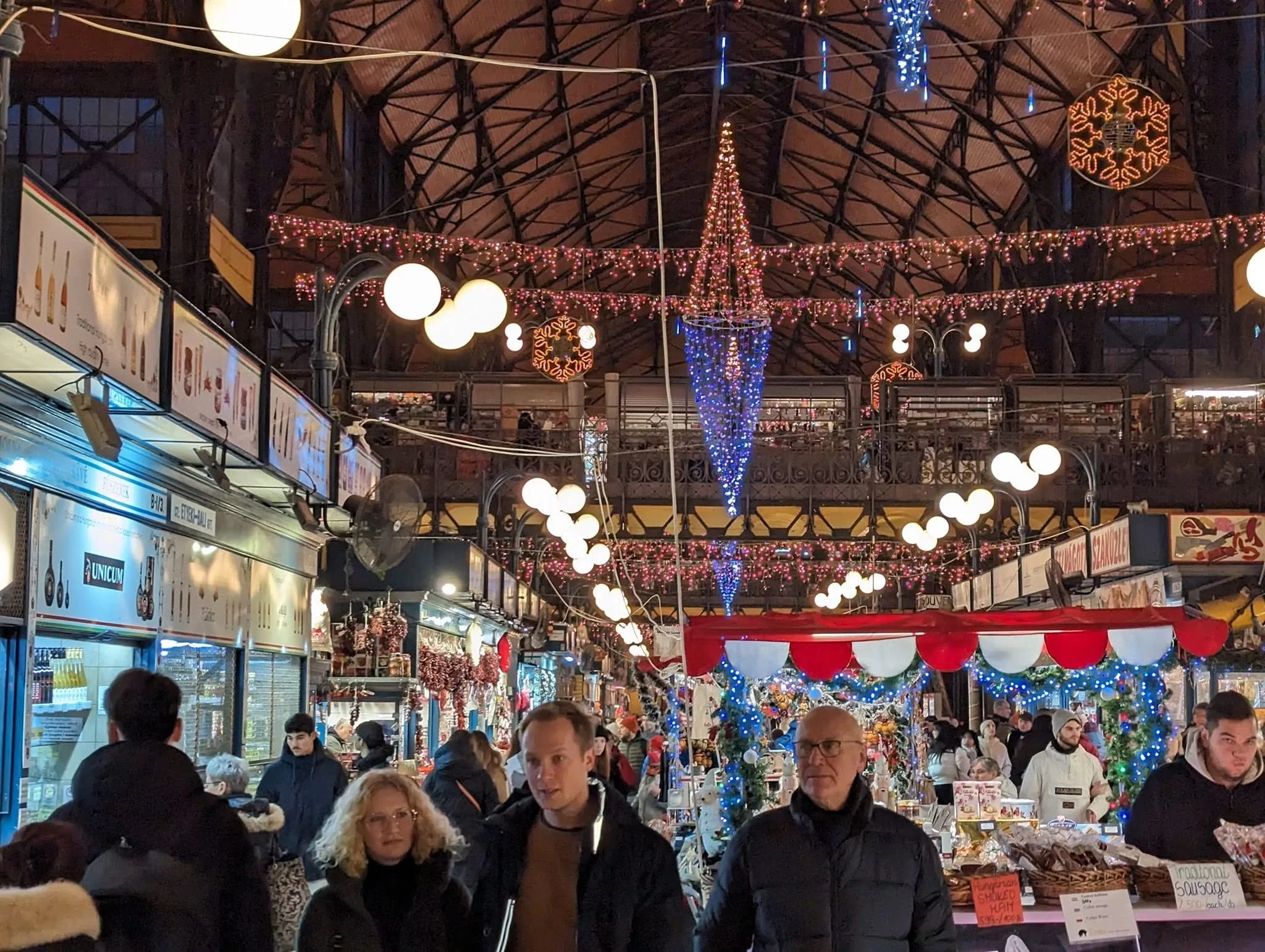 Budapest's Great Market Hall decorated with Christmas lights and festive ornaments, with shoppers browsing market stalls during December 2023.