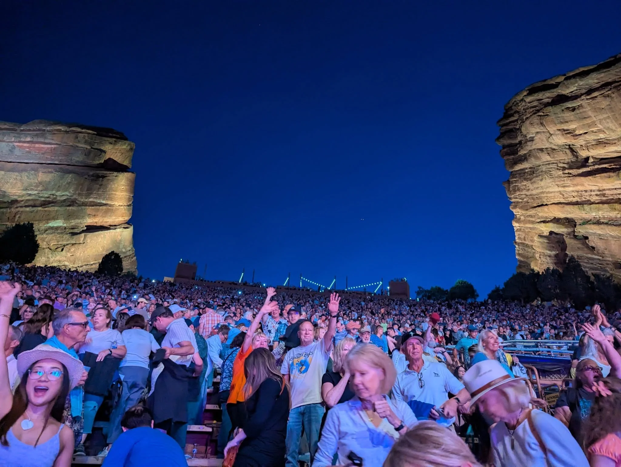 Concert audience at Red Rocks Amphitheatre beneath a deep blue evening sky, framed by two towering sandstone formations.