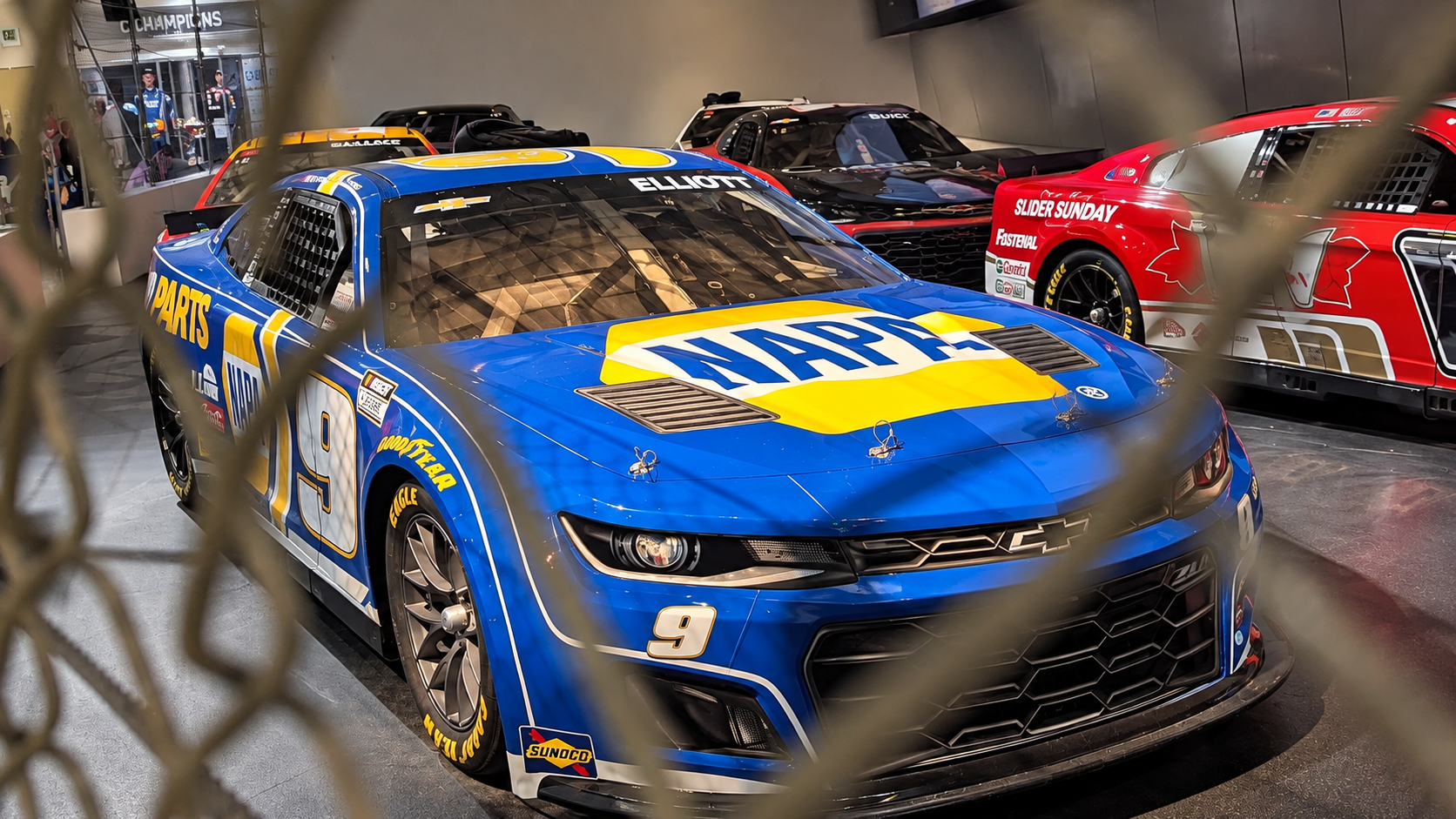 Blue and yellow No. 9 NAPA Chevrolet race car displayed behind fencing at the NASCAR Hall of Fame.