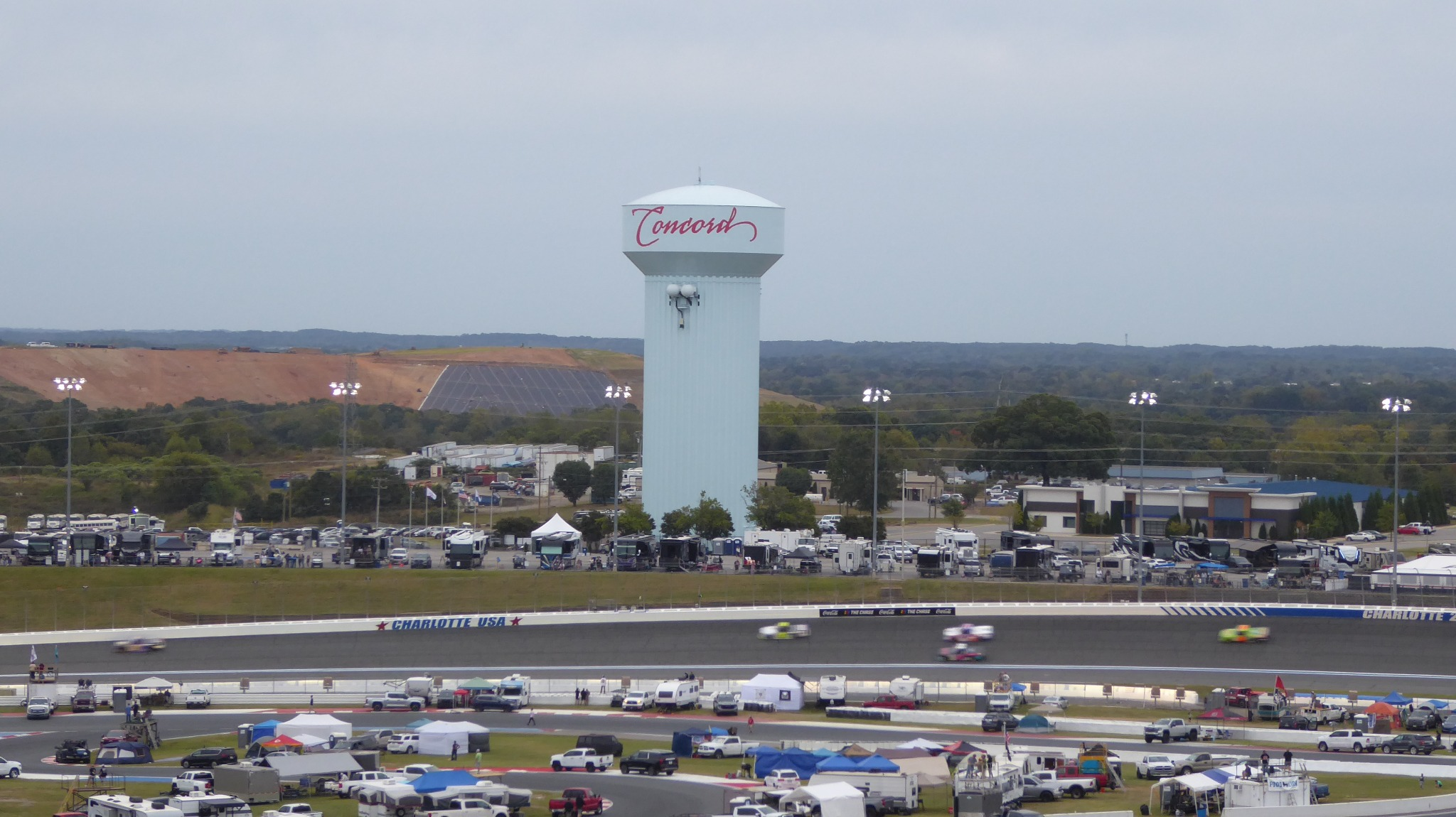 NASCAR cars racing at Charlotte Motor Speedway with the Concord water tower and infield camping area beyond the track.