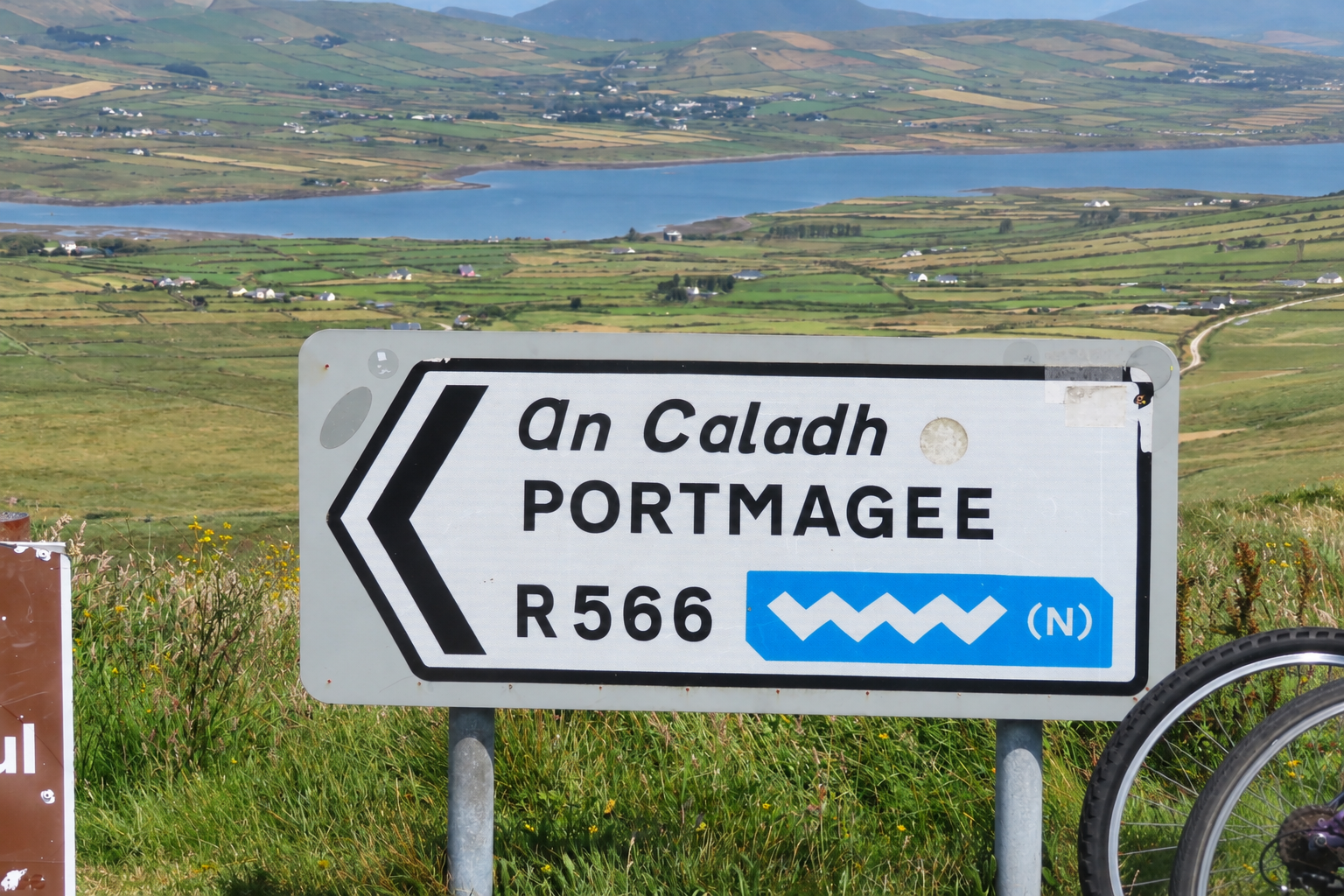 Road sign for Portmagee on Ireland's R566 with green countryside and water in the background, illustrating why road type and drive time matter when planning an Ireland road trip.