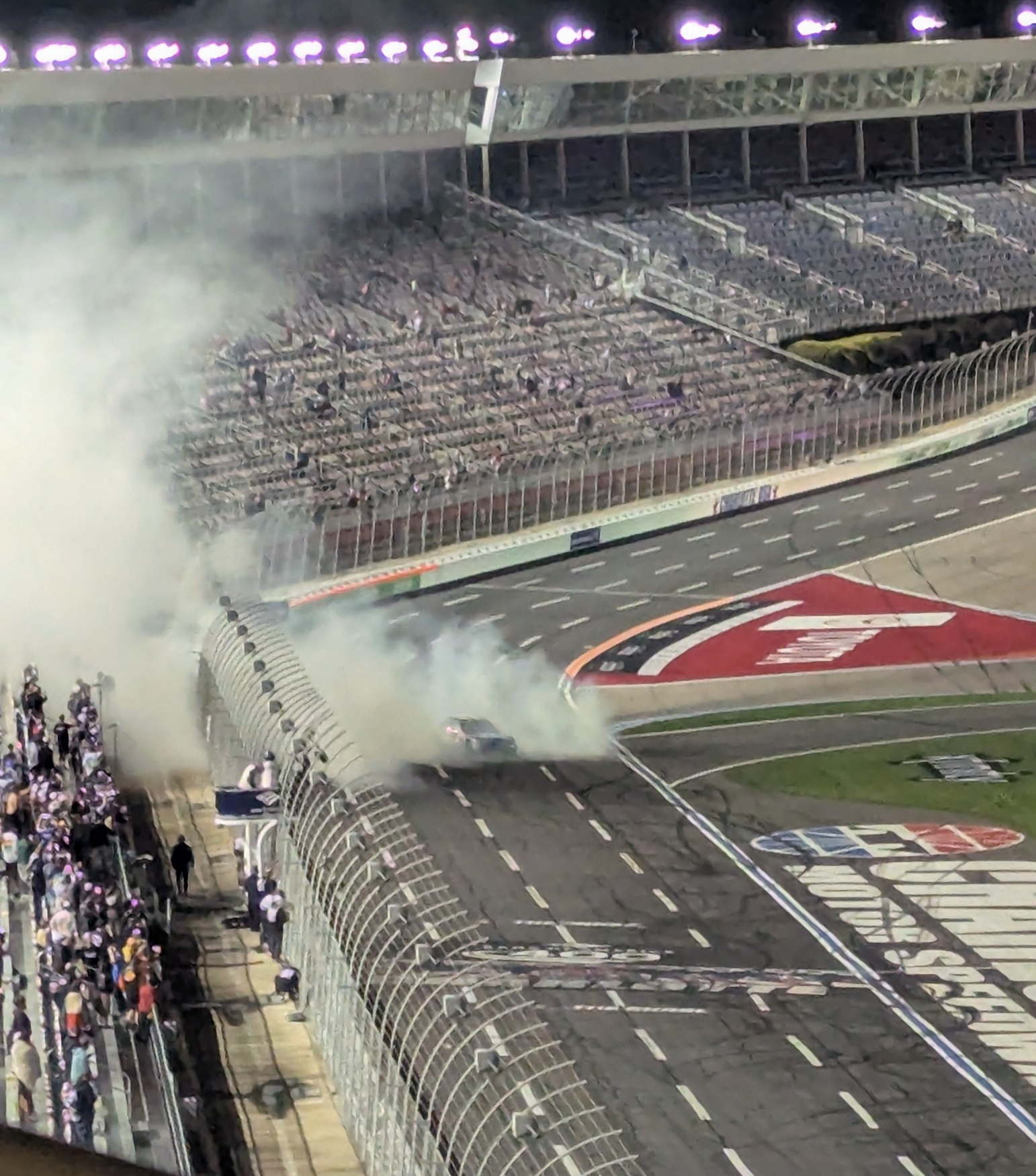NASCAR race car performing a smoky nighttime burnout at Charlotte Motor Speedway as fans watch from the grandstands.
